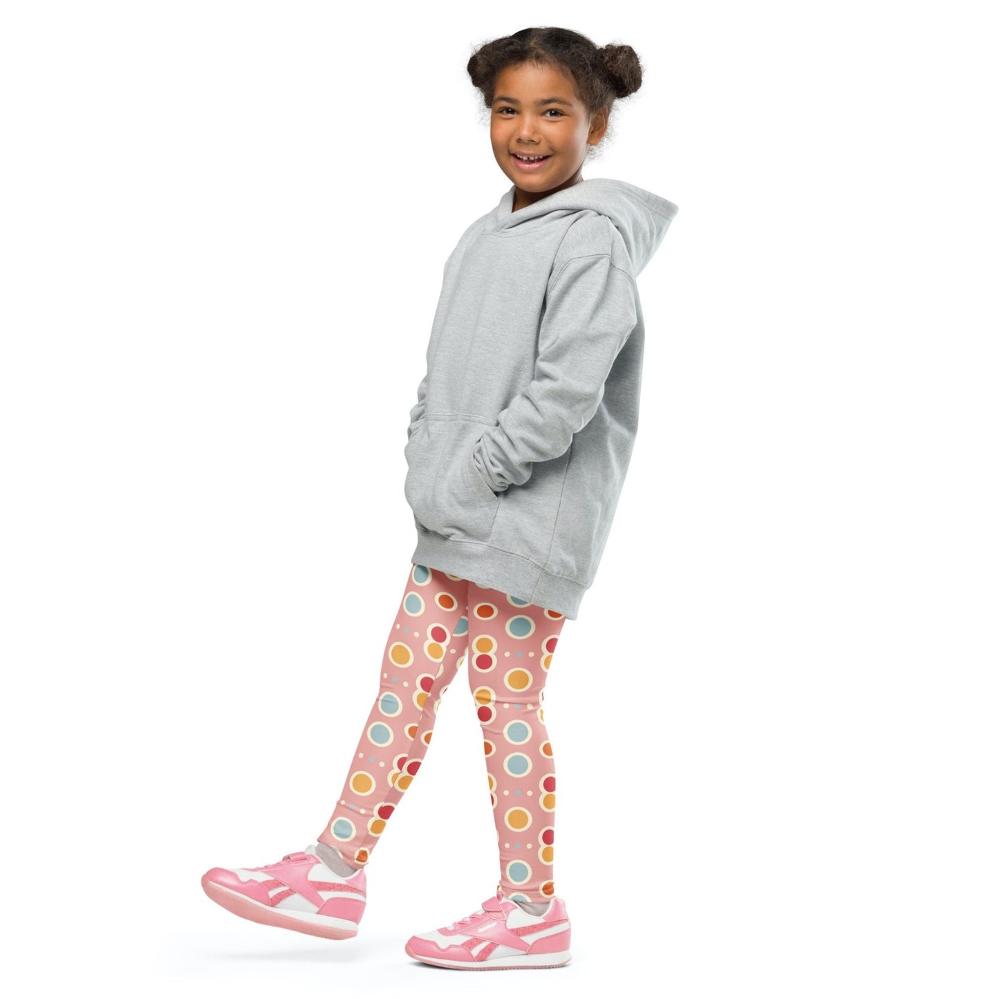 Front left view of a smiling black girl whose hair is bound into buns wearing a grey sweater, and leggings which features a playful pattern of pastel and white circles on a pink background. The girl is standing against a white background.