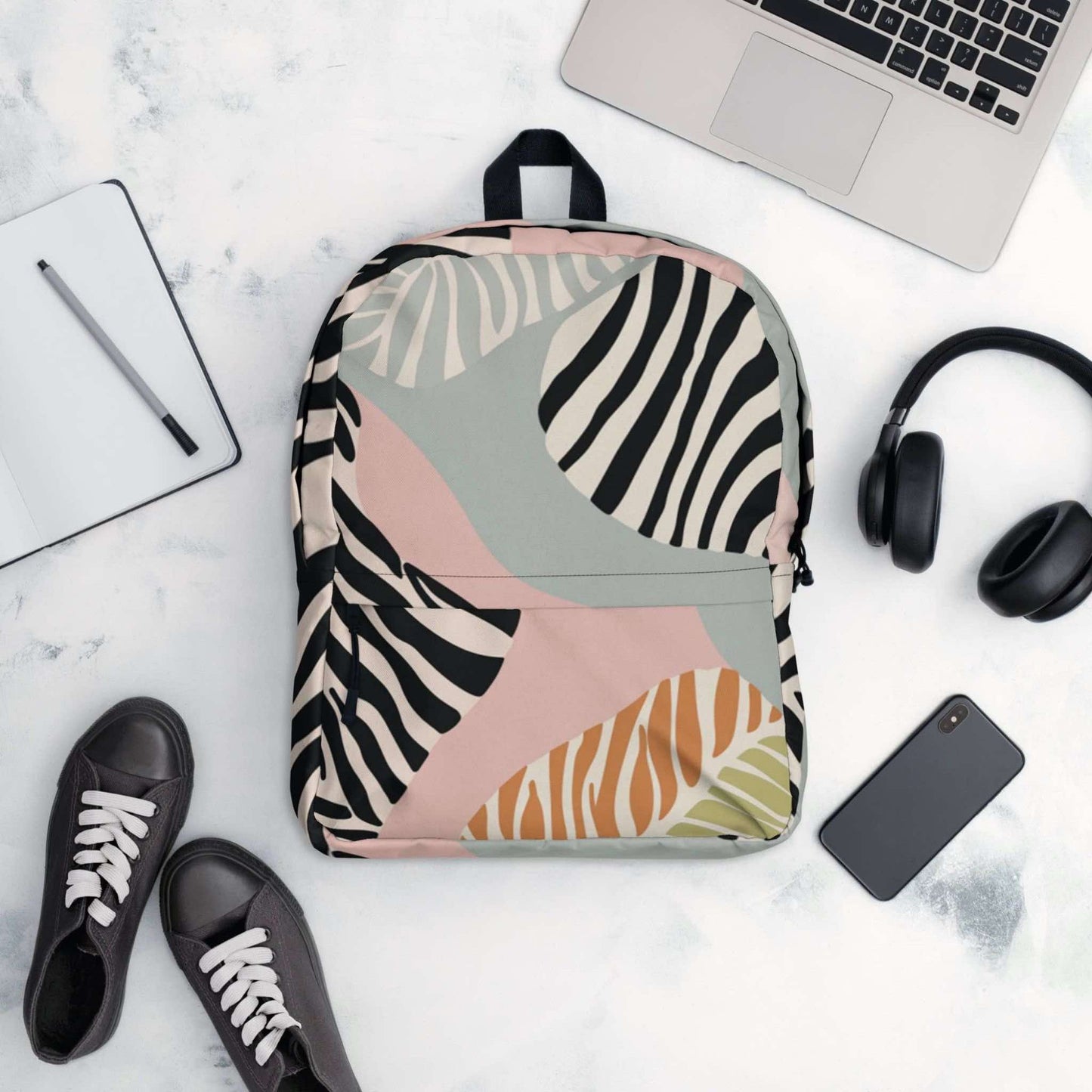 A flatlay of a zebra print backpack, surrounded by items it could conain, like a laptop, handphone, headphone, ontepad and pen, and even shoes.