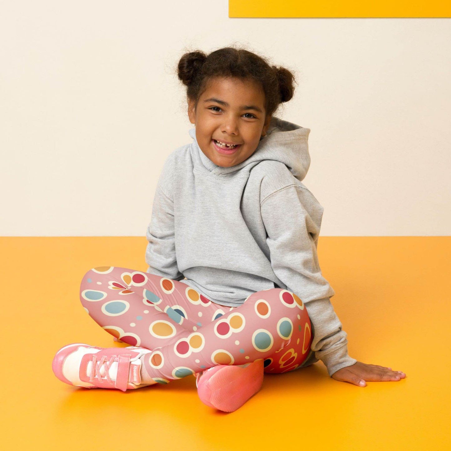 Front view of a smiling black girl whose hair is bound into buns wearing a grey sweater, and leggings which features a playful pattern of pastel and white circles on a pink background. The girl is sitting cross-legged on a yellow floor, against a white and yelllow wall behind her.