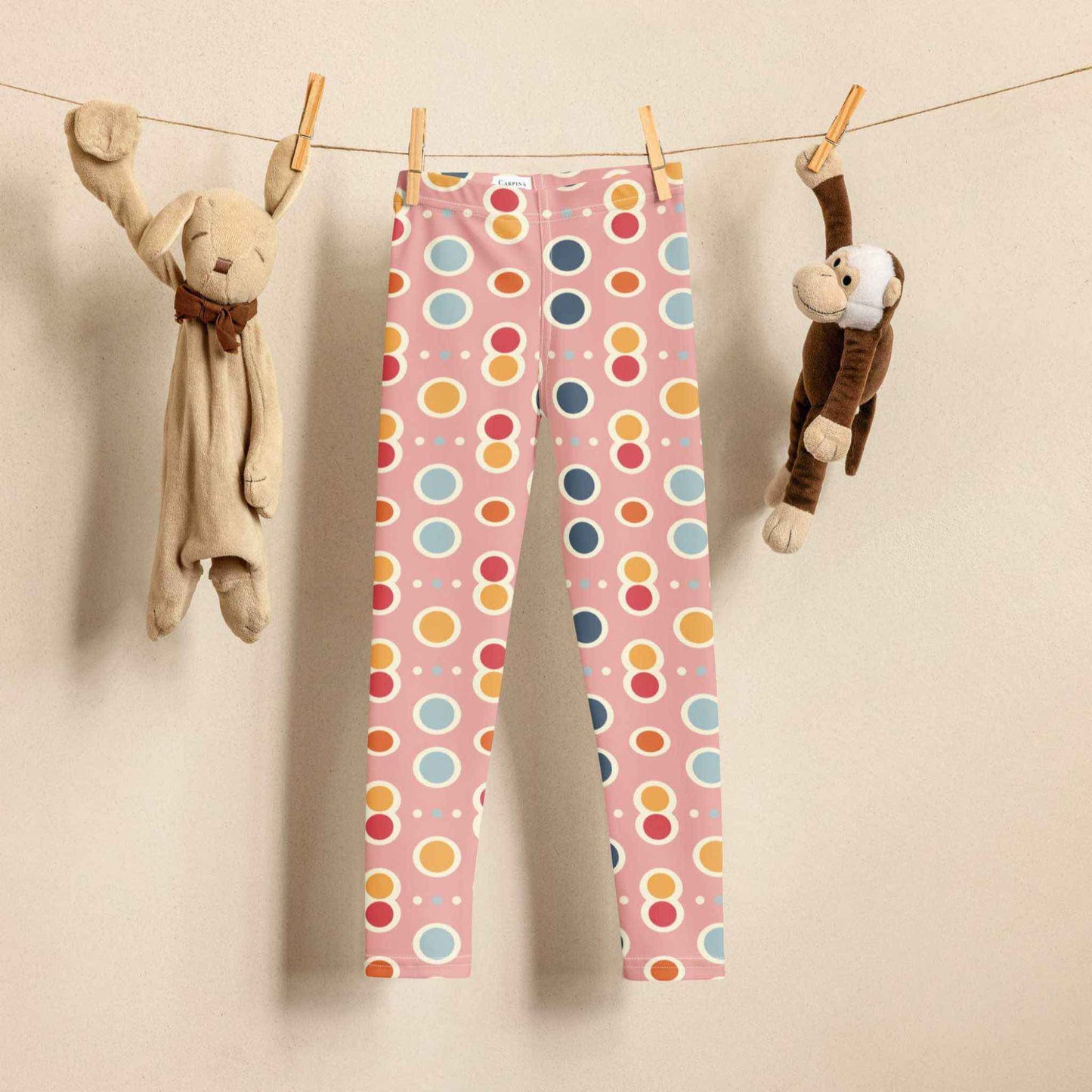 A pair of leggings which features a playful pattern of pastel and white circles on a pink background is hung on a laundry line. A toy rabbit and toy monkey are hung on the same laundry line to the left and right of the leggings respectively.