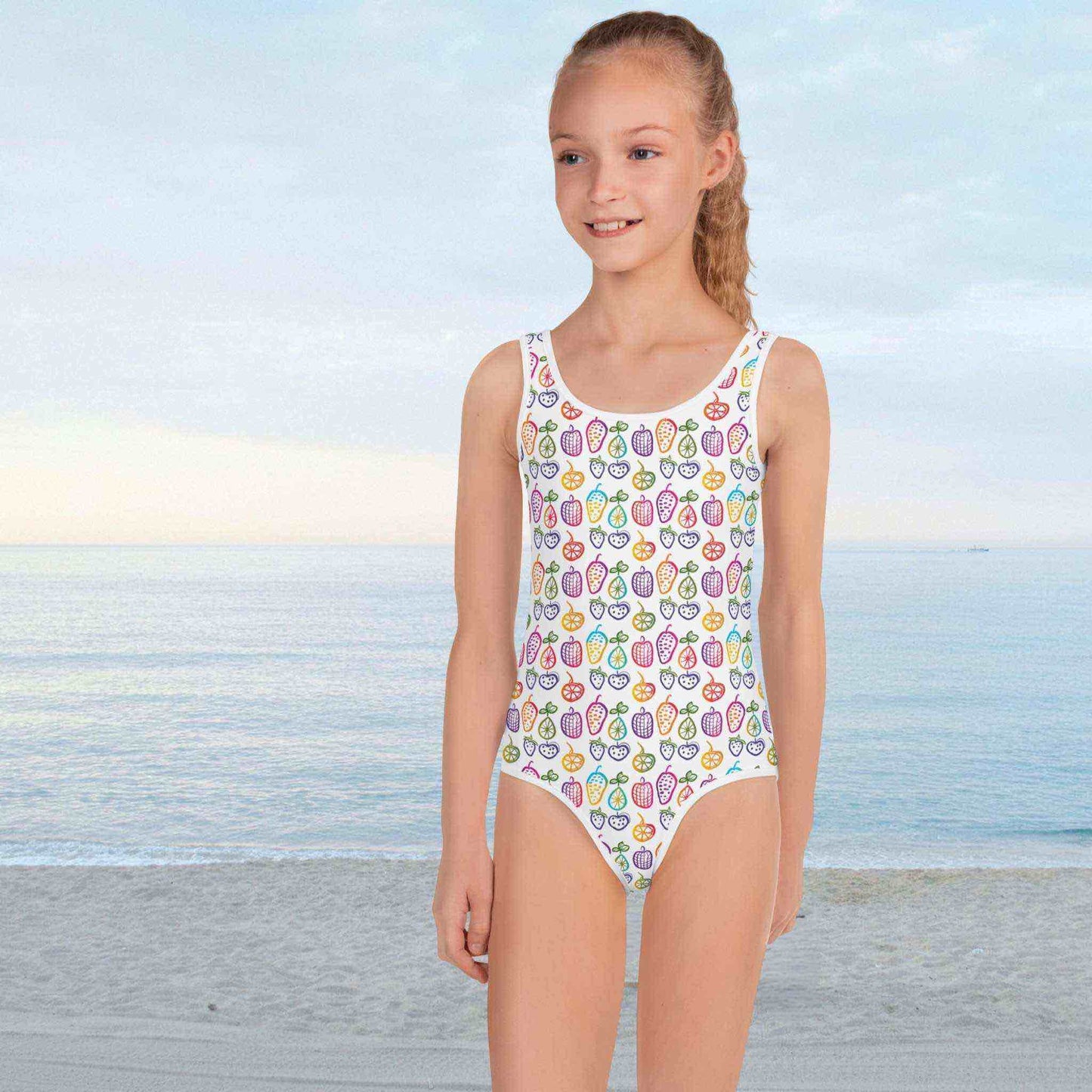 Cute 1 Piece Swimsuits Juniors featuring a cheerful pattern of colorful fruit doodles across a pristine white background, worn by a young girl standing a seas shore.
