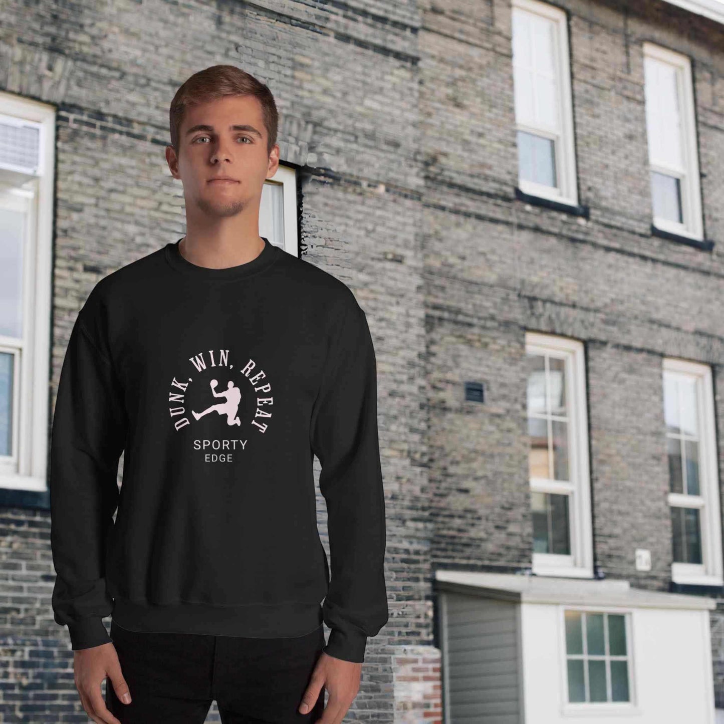 Basketball sweatshirt in black color, with the words "Dunk", "Win", "Repeat" and "Sporty Edge" circulating a graphic of a basketball player in action, worn by a man sanding in front of a classic brick building.