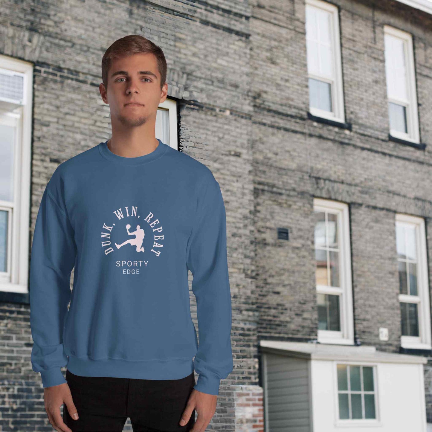 Basketball sweatshirt in indigo blue color, with the words "Dunk", "Win", "Repeat" and "Sporty Edge" circulating a graphic of a basketball player in action, worn by a man sanding in front of a classic brick building.