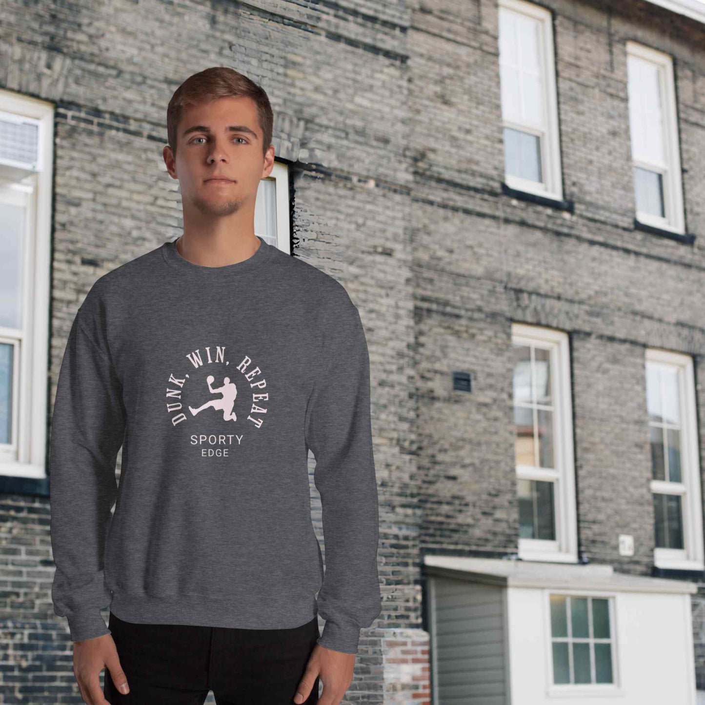 Basketball sweatshirt in dark heather color, with the words "Dunk", "Win", "Repeat" and "Sporty Edge" circulating a graphic of a basketball player in action, worn by a man sanding in front of a classic brick building.