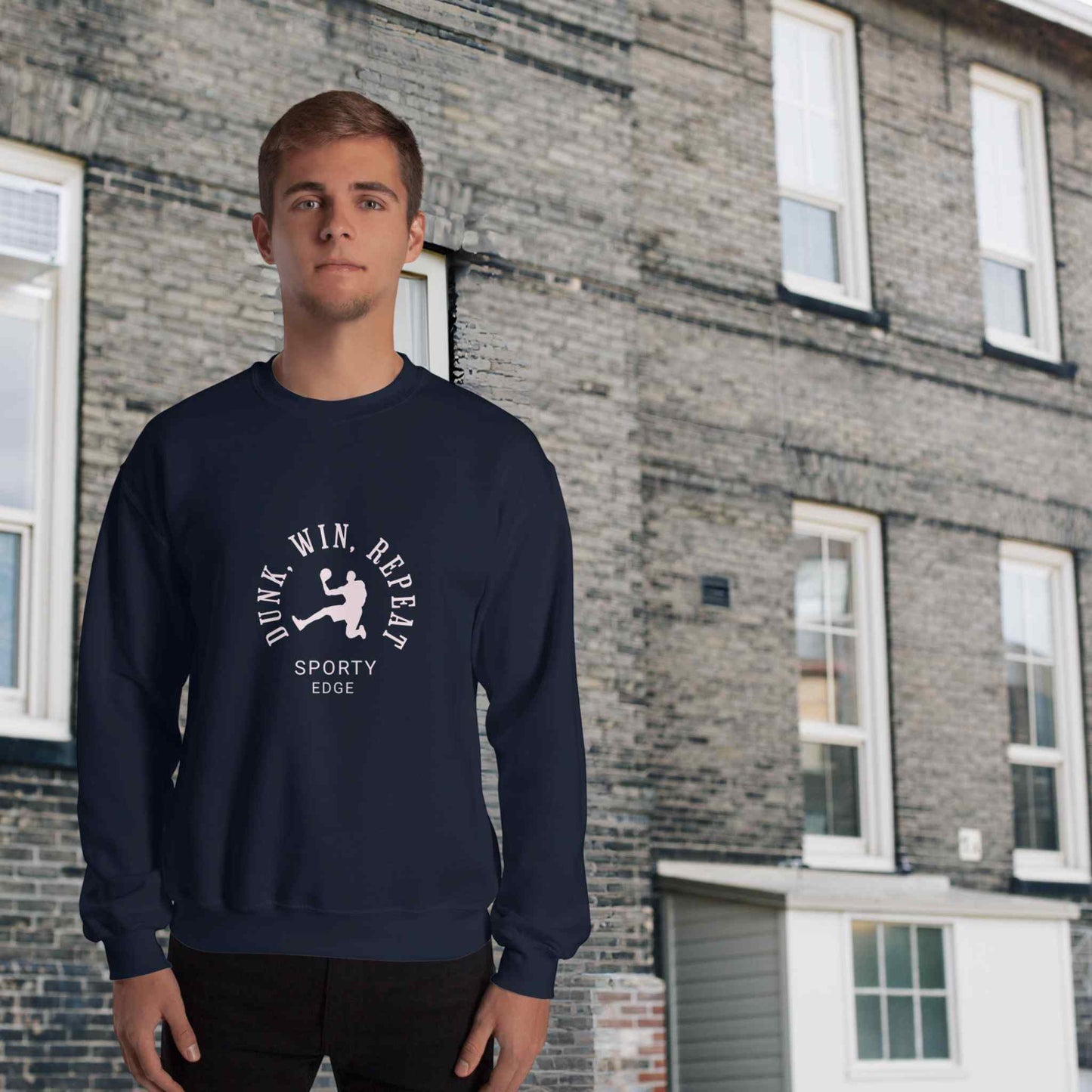 Basketball sweatshirt in navy color, with the words "Dunk", "Win", "Repeat" and "Sporty Edge" circulating a graphic of a basketball player in action, worn by a man sanding in front of a classic brick building.