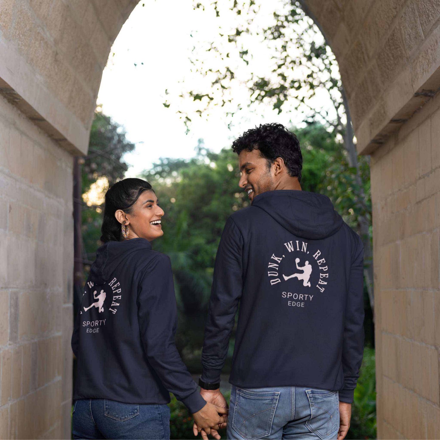 Basketball hoodie, in Navy Blazer color, worn each by a couple, holding hands and smiling at each other, standing under an arch with their backs to the camera. The words on the hoodie, "Dunk, Win, Repeat" and "Sporty Edge" circle around a motif of a basketball player in action.