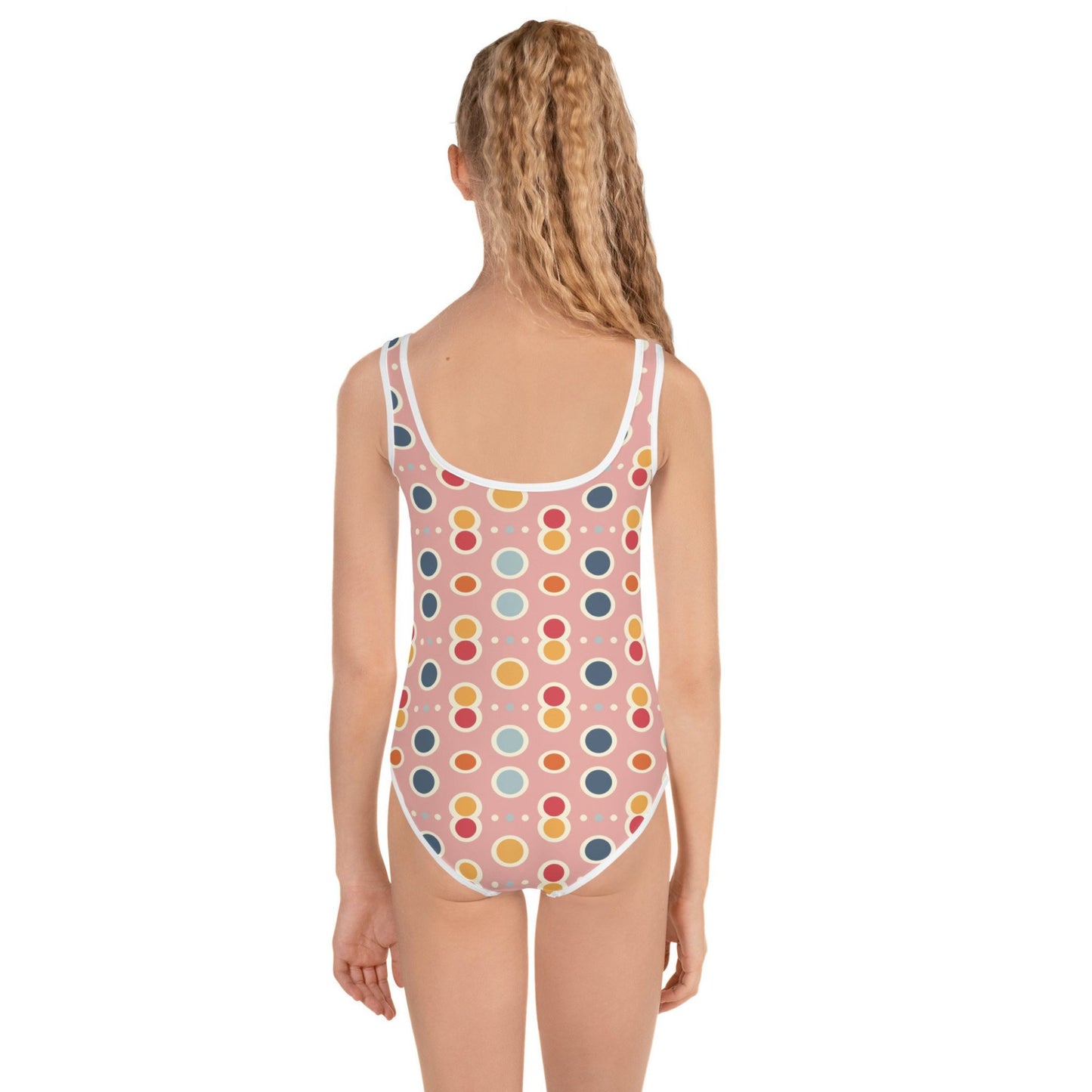 Adorable infant one piece swimsuit, which is also for kids, junior features a playful pattern of pastel pops and white outlines on a pink background, worn by a young girl, her back facing the camera. White background.