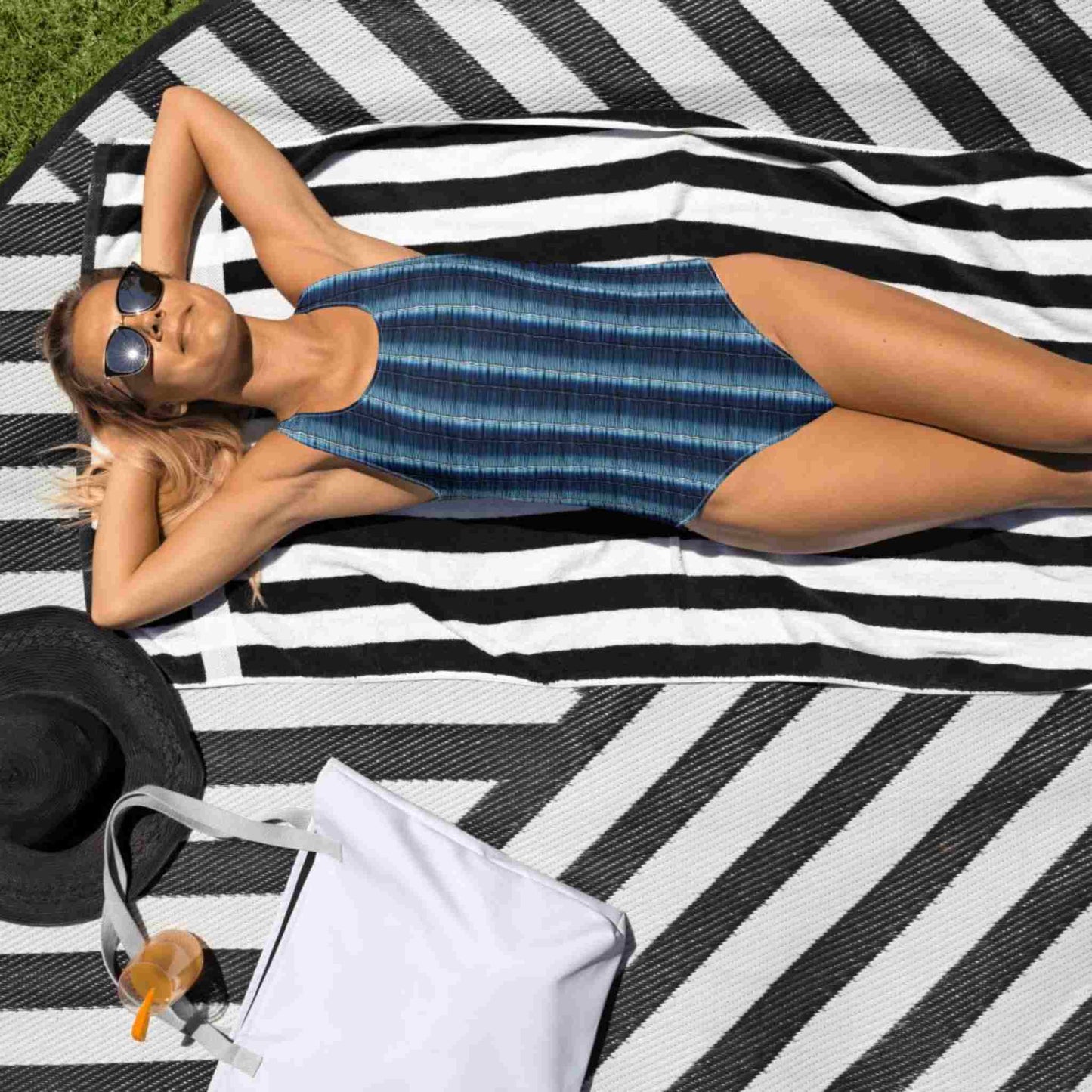 Slimming one-piece swimsuit which features a flattering denim print with vertical stripes, worn by a woman with sunglasses lying on a beach mat in the sun.