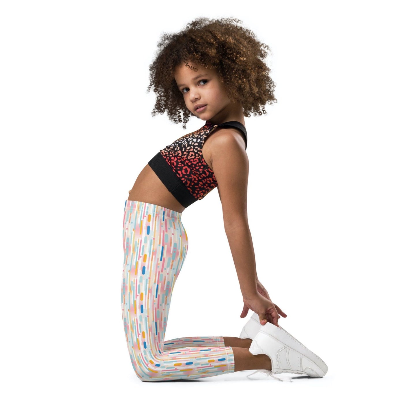 Kid and roddler leggings which features playful candy colored lines in a white background, worn by a young girl. The girl kneels and stretches backwards, in a side view to camera, her face lited left towards the camera. White background.