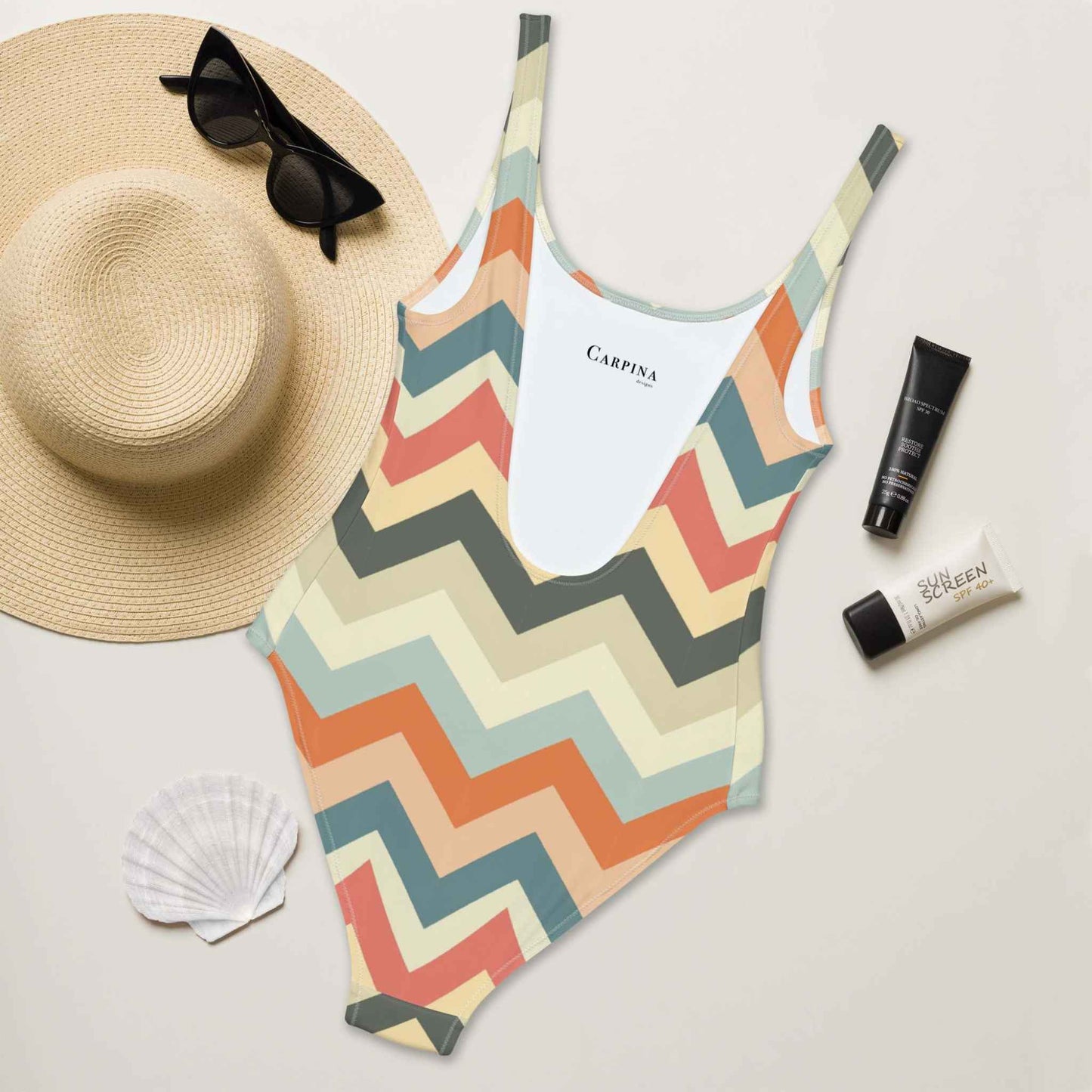 A flat lay of a one piece swimsuit, with rear side facing up, designed in a chic zig zag pattern in pastel shades. A hat, sunglasses, seashell and tubes of sun screen creams laying next to it. The Carpina Designs logo printed on the inside of the swimsuit is visible.