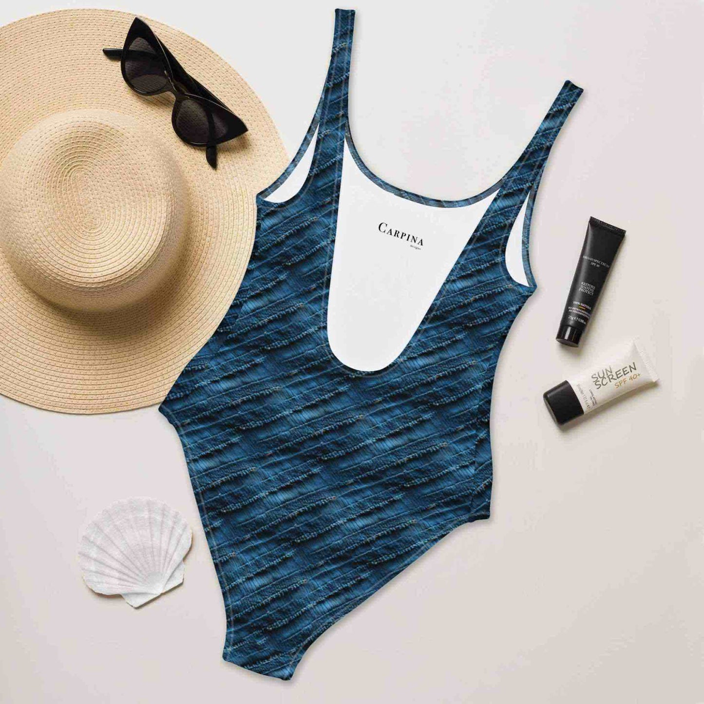 Flatlay of a blue pattern swimsuit (one piece)Blue pattern swimsuit (one-piece) which features a chic denim print with subtle stripes, rear side facing up, creating a flattering, slimming effect, surrounded by a straw hat, sunglasses, sea shore and sunscreen lotions.