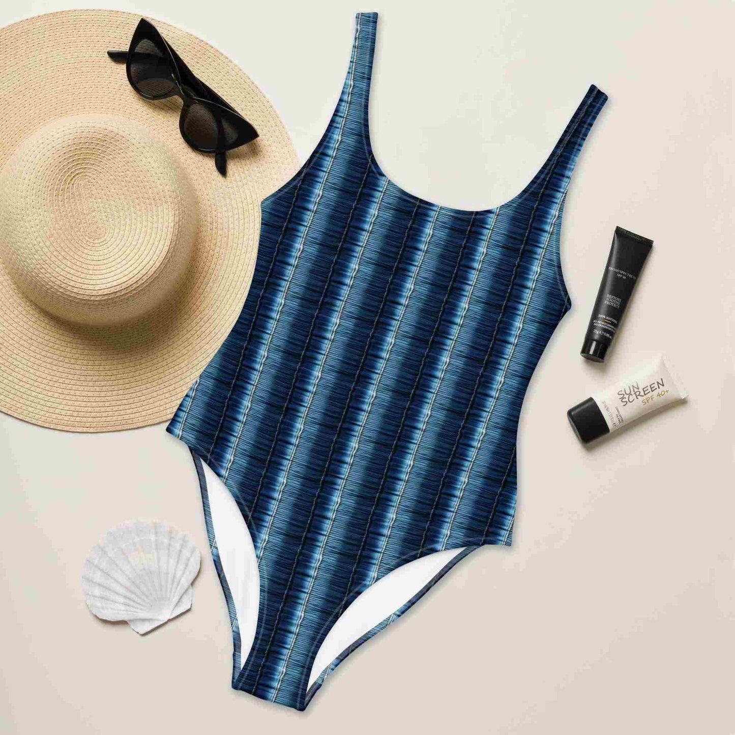 Flatlay of a slimming one-piece swimsuit which features a flattering denim print with vertical stripes, front side facing up, surrounded by a straw hat, sunglasses, sea shell and sunscreen lotions.