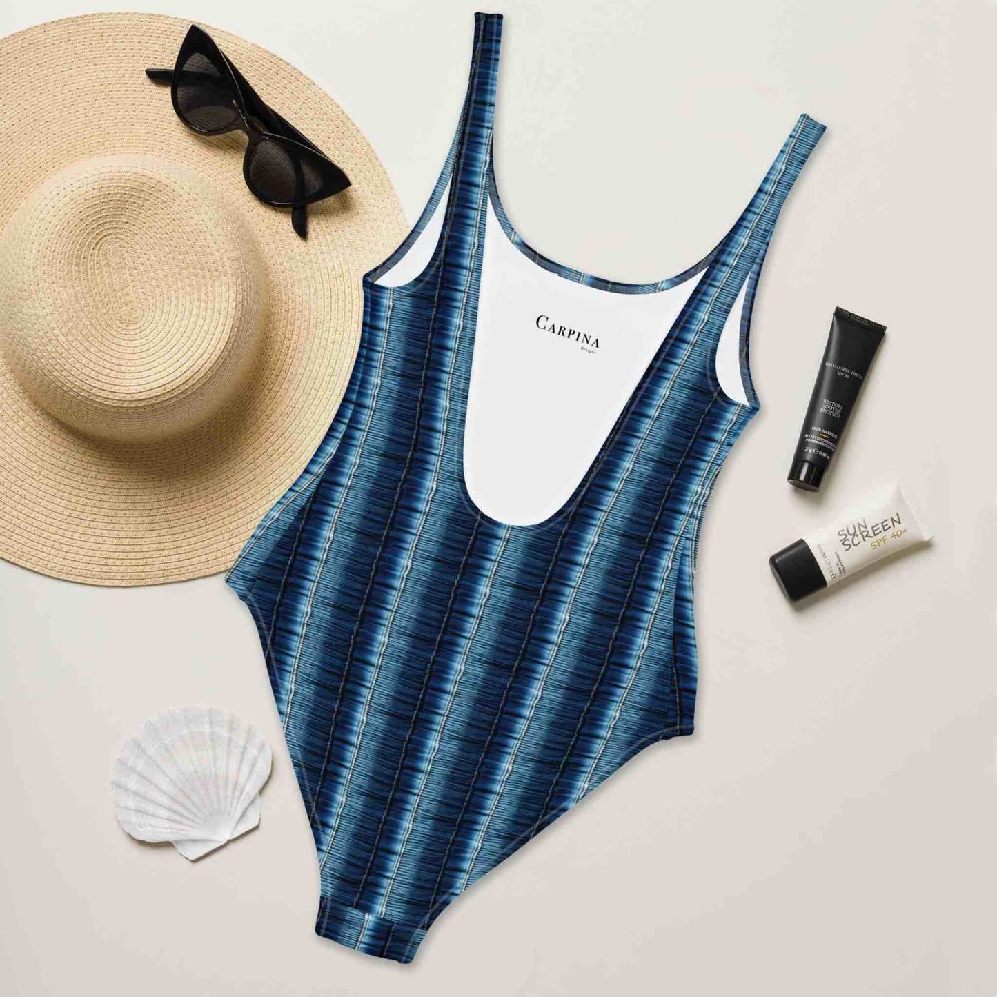 Flatlay of a slimming one-piece swimsuit which features a flattering denim print with vertical stripes, rear side facing up, surrounded by a straw hat, sunglasses, sea shell and sunscreen lotions.