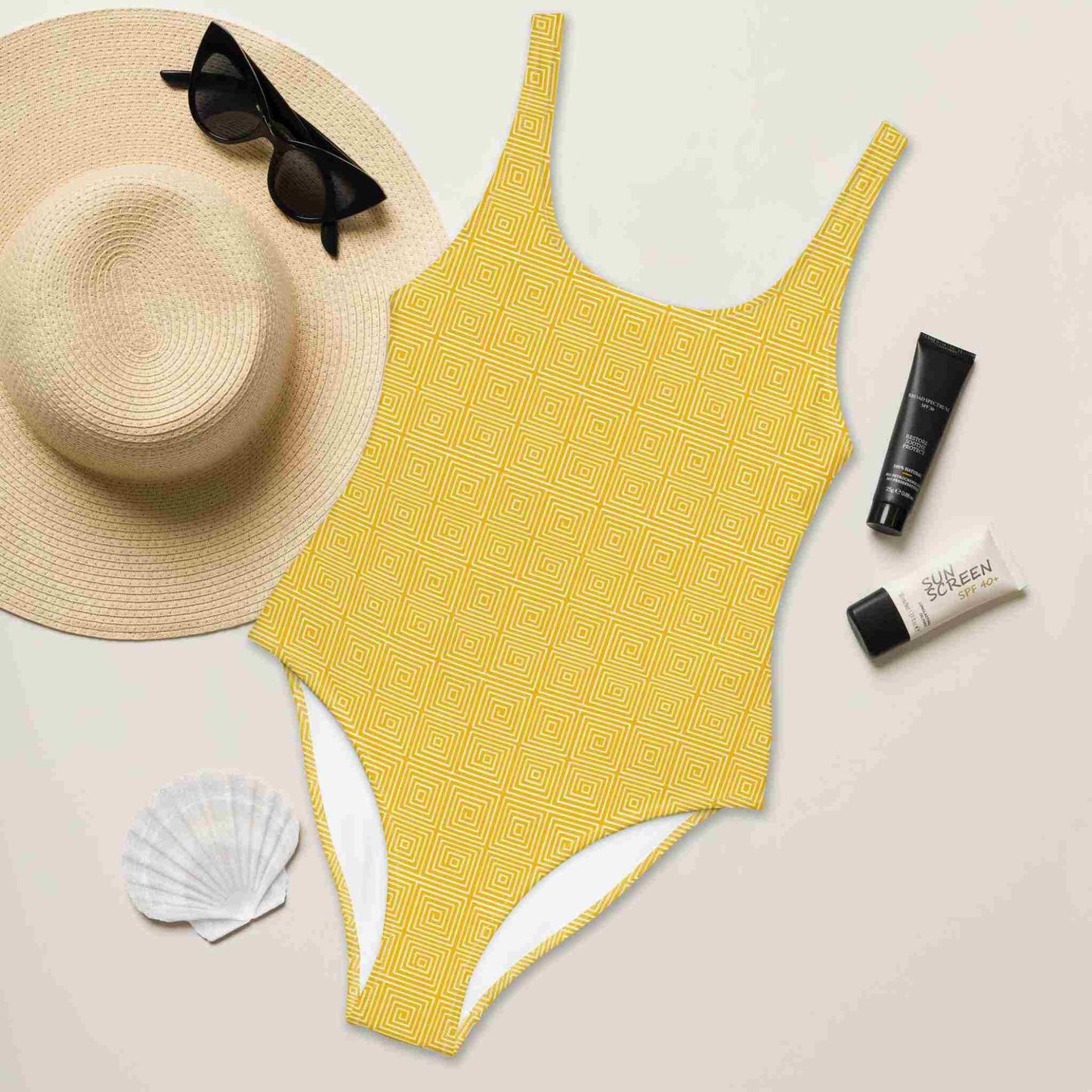 Flatlay of a yellow bathing suit one piece which features a yellow op art illusion pattern, front facing up, surrounded by a hat, sunglass, sea shell and sunshade lotions.