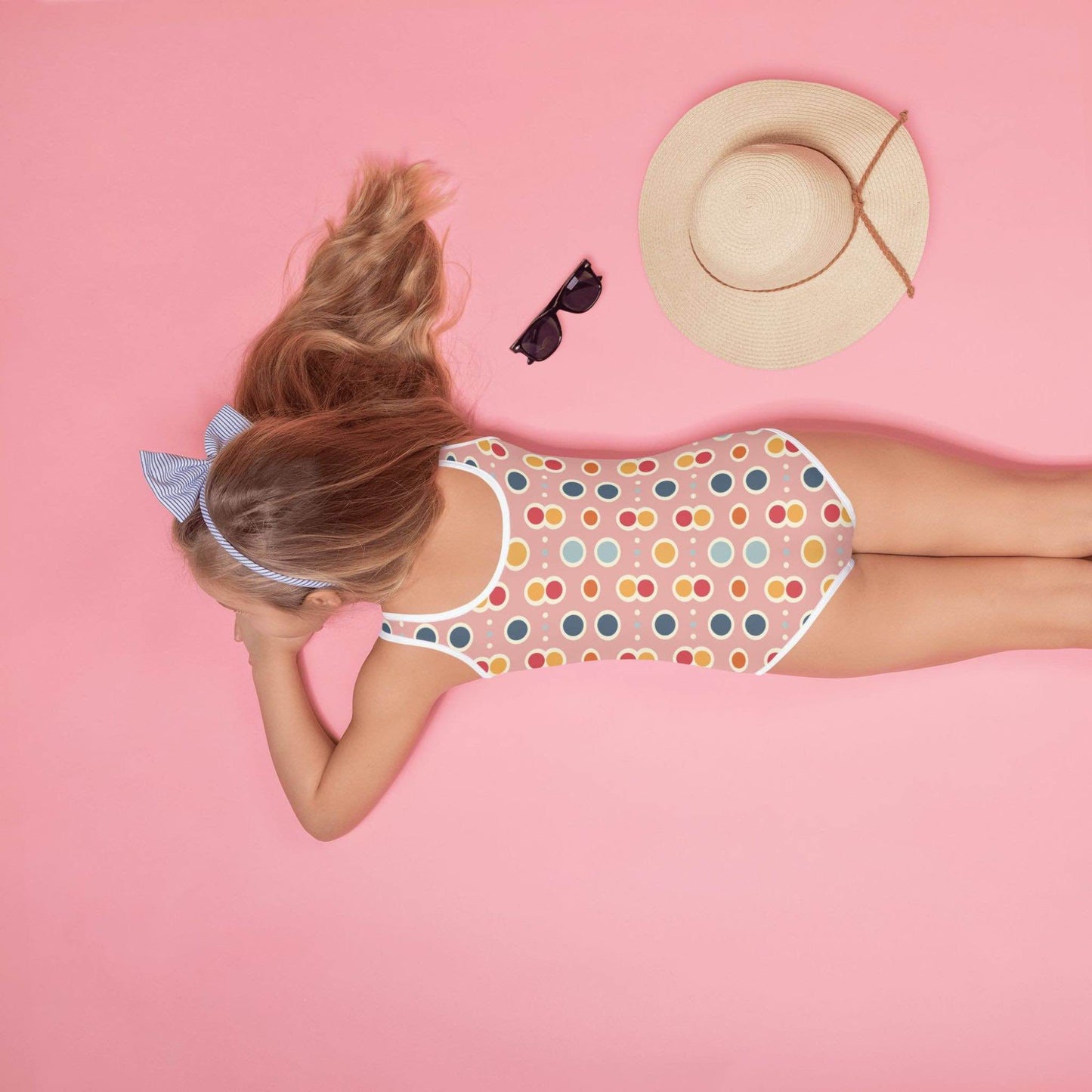 Adorable infant one piece swimsuit, which is also for kids, junior features a playful pattern of pastel pops and white outlines on a pink background, worn by a little girl, lying face down on a pink background. A straw hat and sunglasses are on her side.