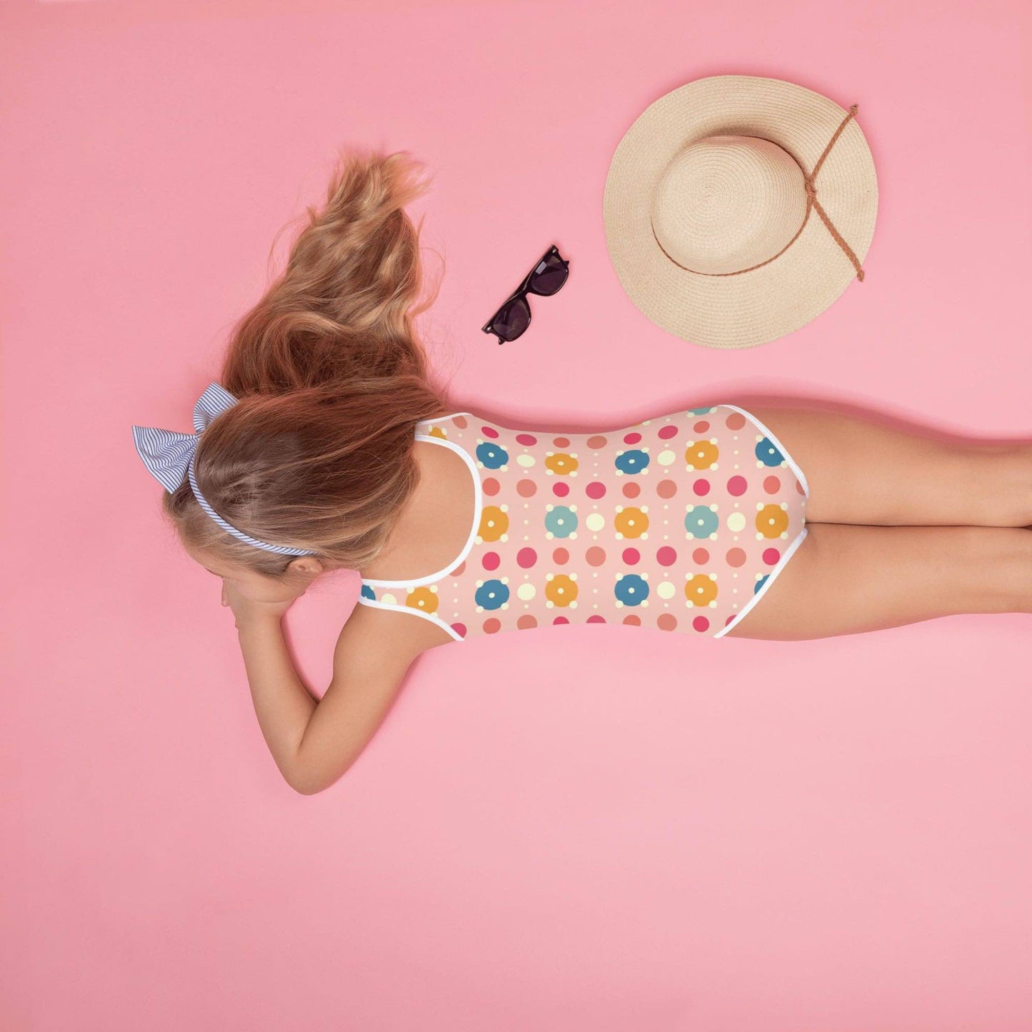 Cute infant swimsuit, suitable for kids in general, features a playful pattern of pastel and white circles on a pink background, worn by a little girl, lying face down on a pink background, surrounded by a sunglass and straw hat.