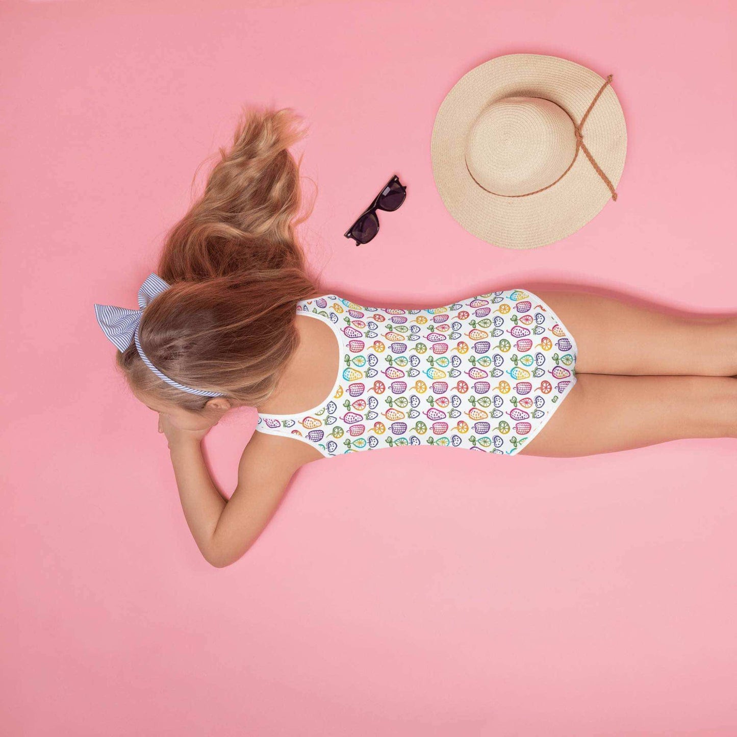 Cute 1 Piece Swimsuits Juniors featuring a cheerful pattern of colorful fruit doodles across a pristine white background, worn by a young kid. She is lying face down on a pink background with a straw hat beside her..