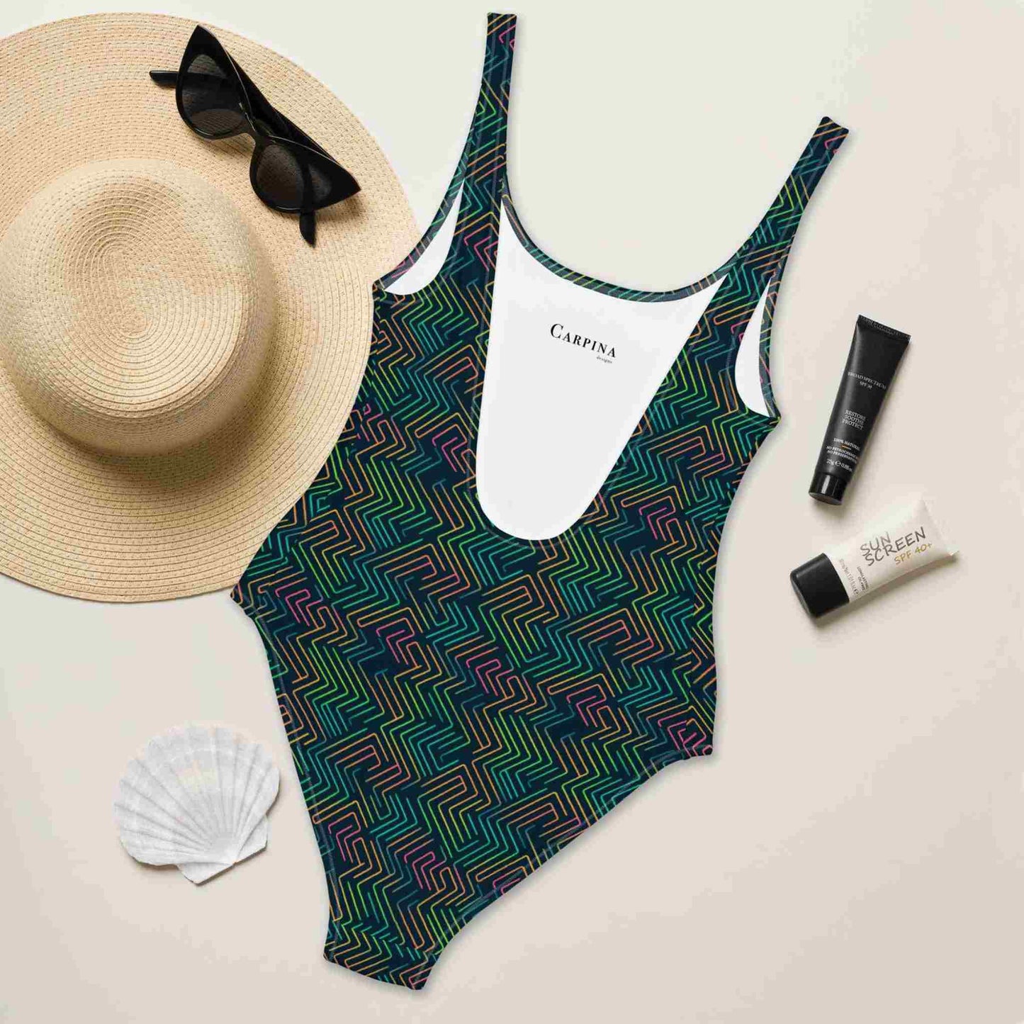 Flat lay of aDark green one-piece swimsuit featuring a chic dark green neon pattern, rear side facing up, surrounded by a straw hat, sunglasses, sea shell and sunscreen lotions.