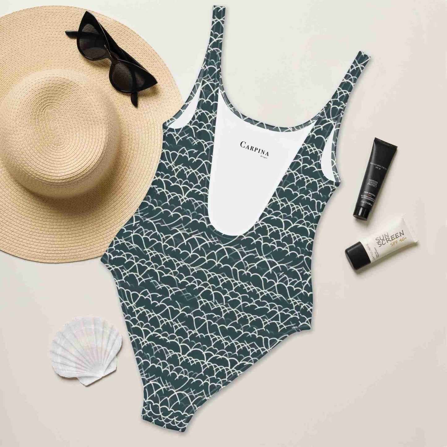 Flat lay of a blue and white pattern swimsuit (one piece) which features a white geometric white design that dances across a blue background, rear side facing up, surrounded by a hat, sunglass, sea shell and sun lotion.