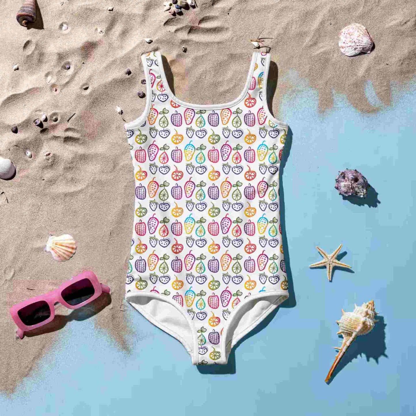 Flatlay of a cute 1 Piece Swimsuits Juniors featuring a cheerful pattern of colorful fruit doodles across a pristine white background, lying with front side up worn by a baby,.on a sandy light blue surface surrounded by a sunglass and sea shells.