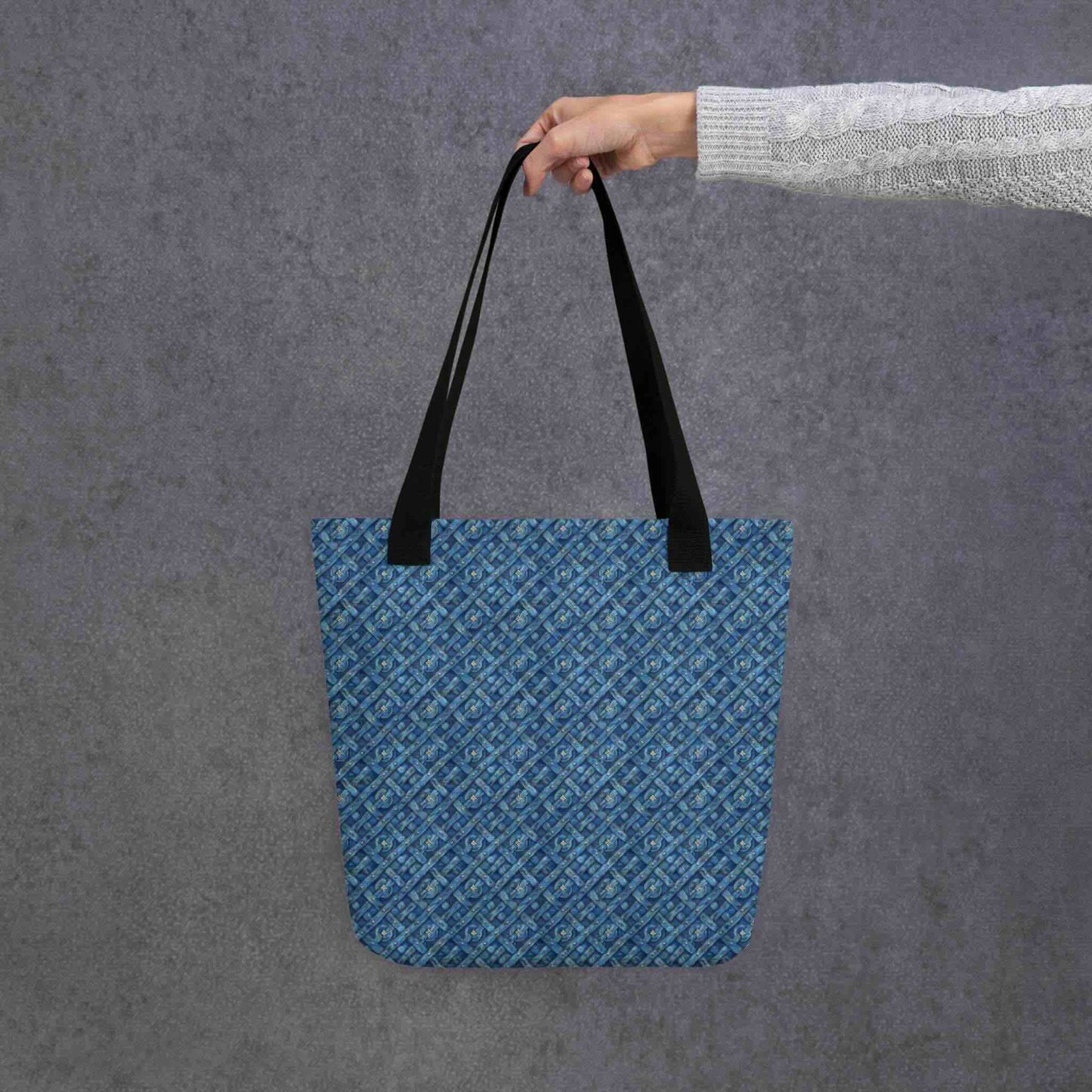 Blue tote bag with black handles, which features a blue Viking inspired denim pattern, held by a lady's hand. Dimension 15x15 inches. Grey background.