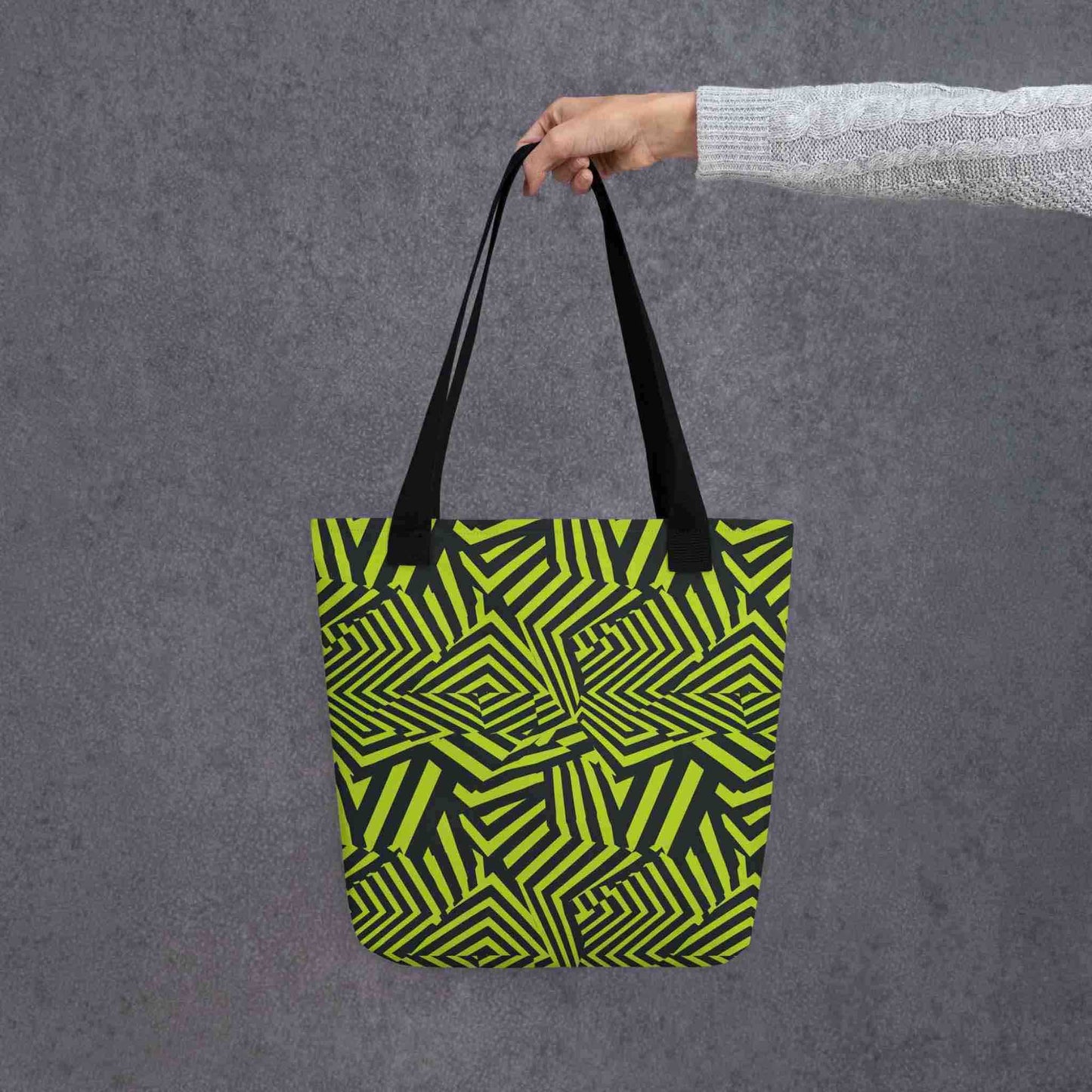 An avant garde tote bag with black handles, which features bold black and green op art illusion, held in a lady's hand. Size is 15x15 inch. Grey background.