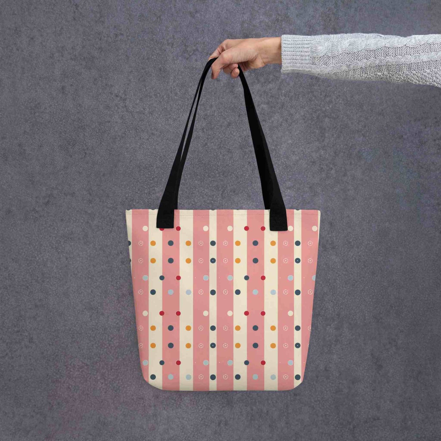 Pink striped tote bag with black handles, which features pink and beige stripes with colourful pokka dots, held by a lady's hand. Dimension 15x15 inches. Grey background.