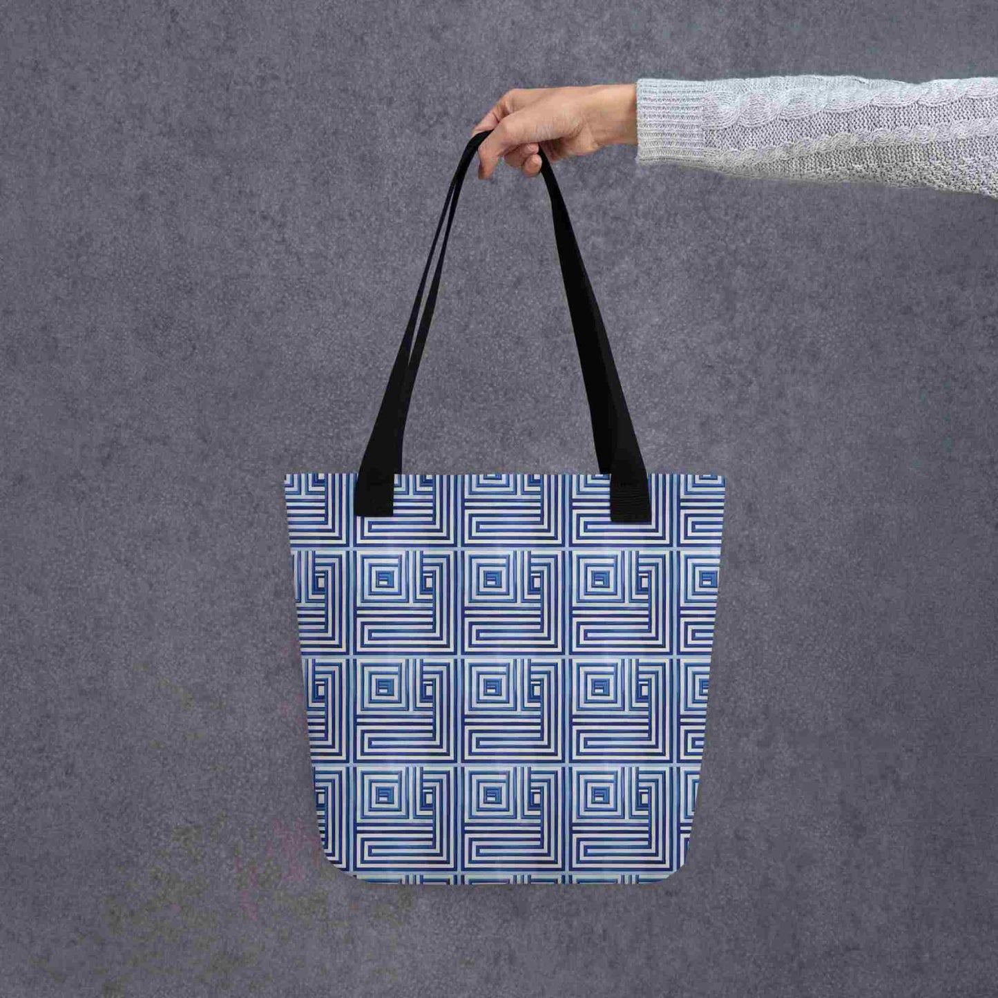 Blue op art tote with black handles, which features a blue op art illusion, held in a lady's hand. Dimension is 15x15 inches. Grey background.