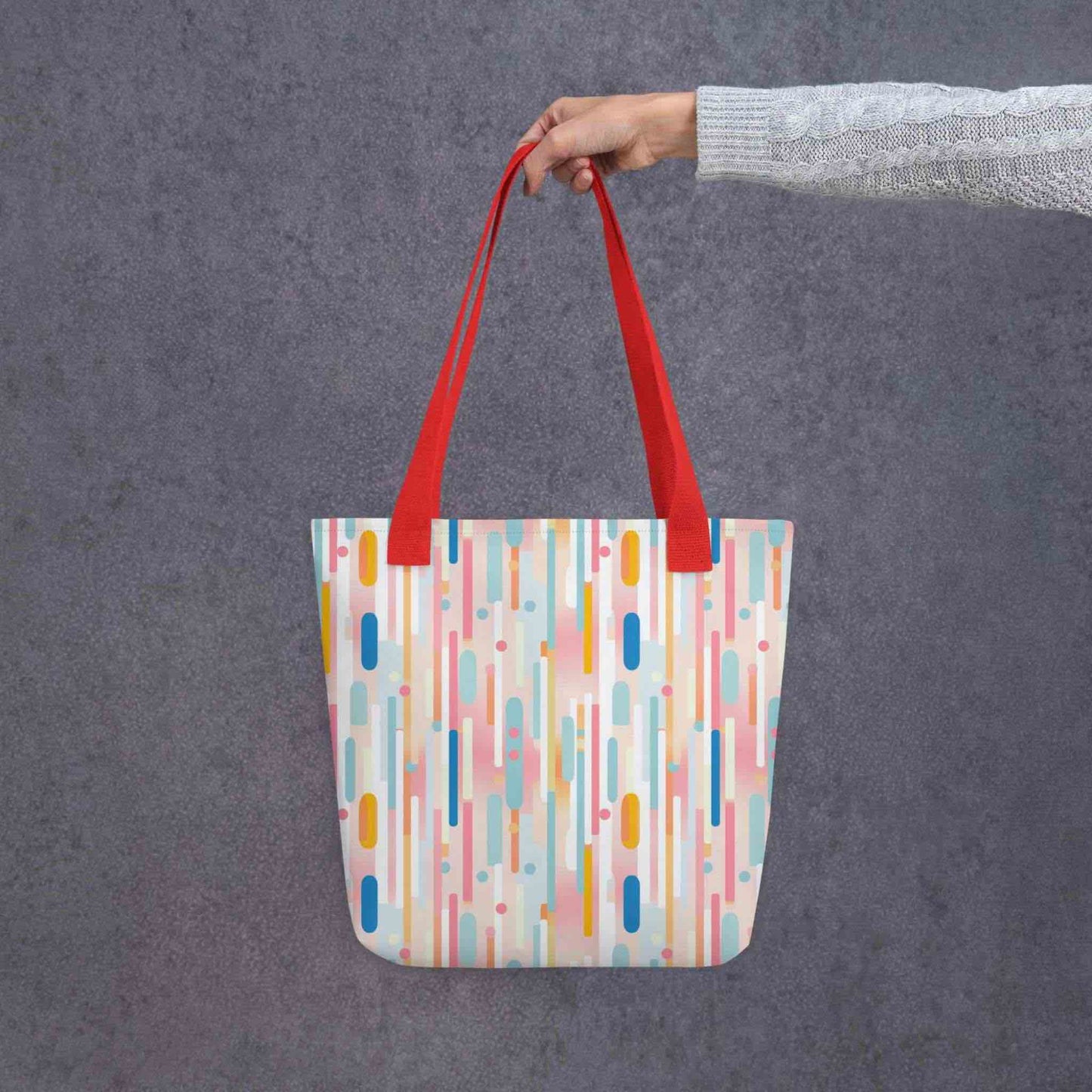 Light pink tote bag with red handles, which design is abstract vertical lines of pastel pink and other pastel shades, held by a lady's hand. Dimension 15x15 inches. Grey background.