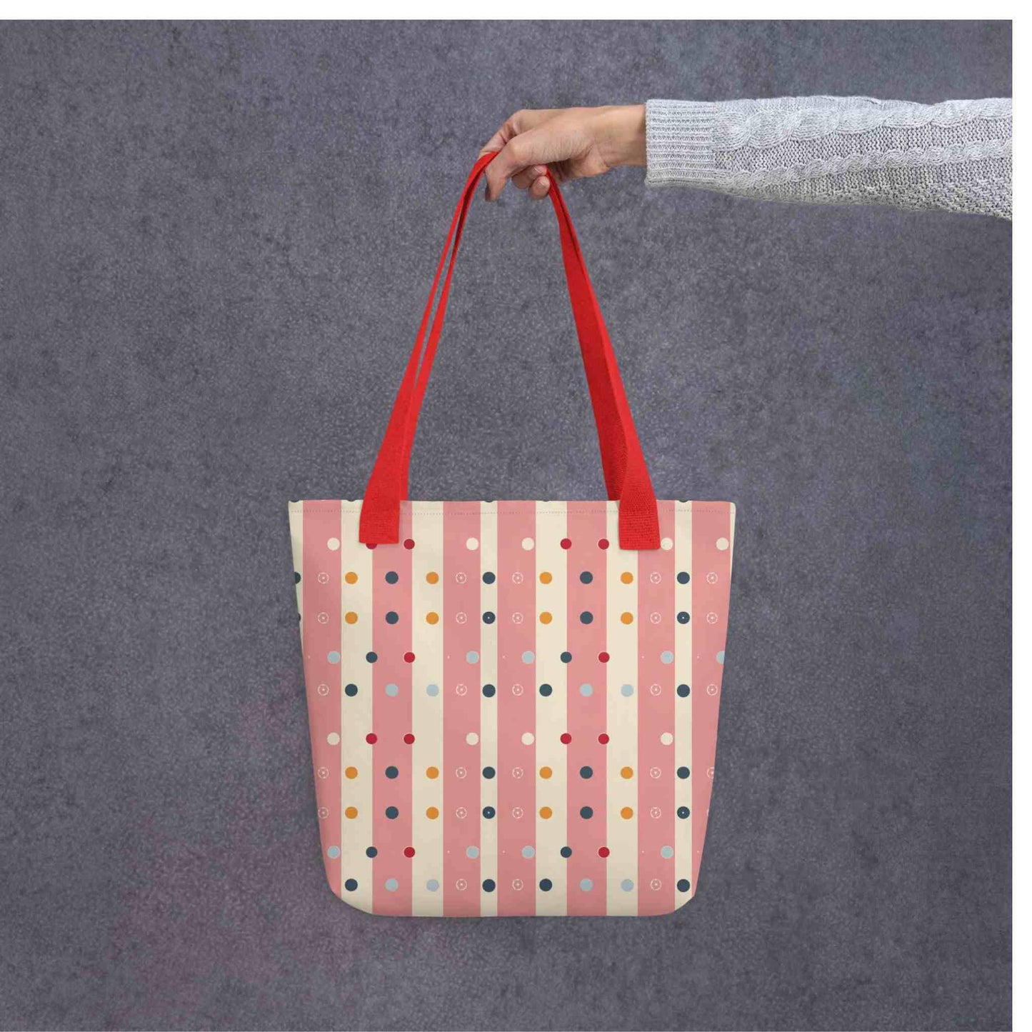 Pink striped tote bag with red handles, which features pink and beige stripes with colourful pokka dots, held by a lady's hand. Grey background.