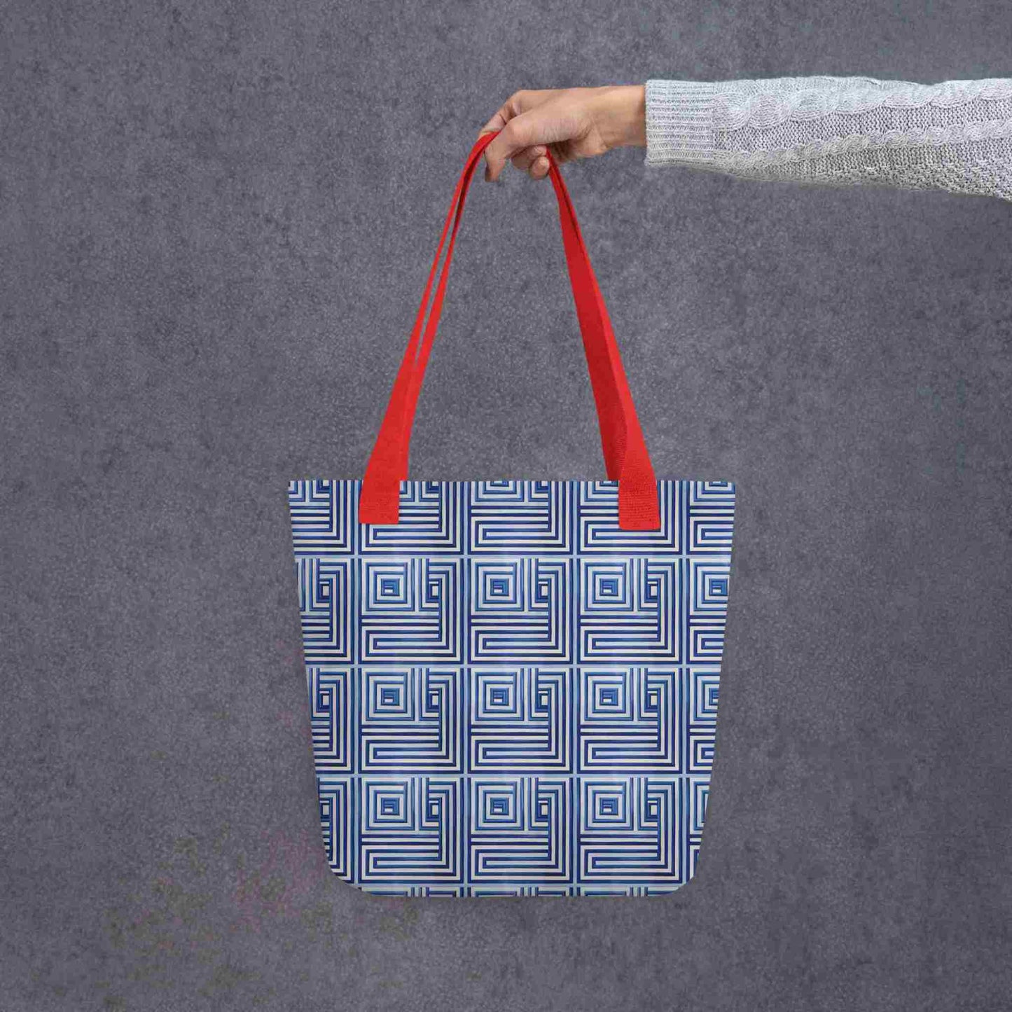 Blue op art tote with red handles, which features a blue op art illusion, held in a lady's hand. Dimension is 15x15 inches. Grey background.