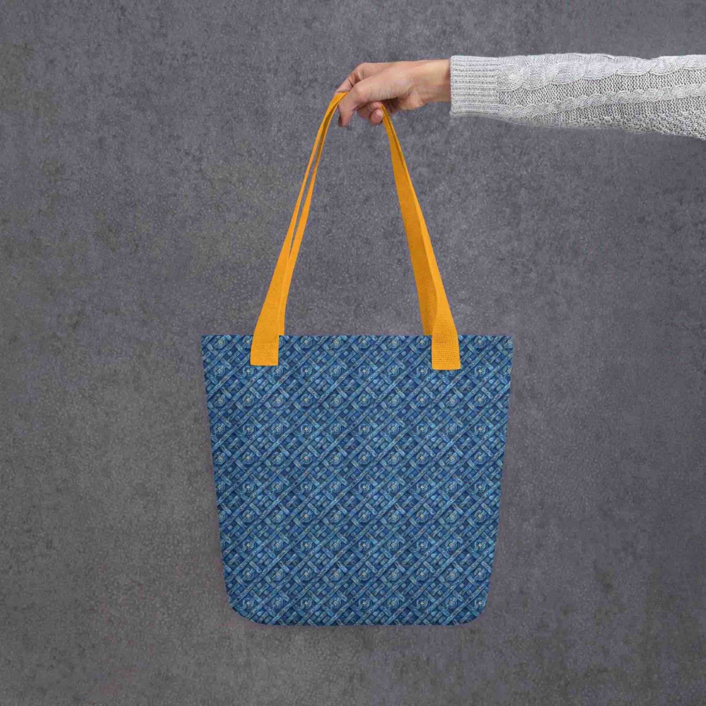 Blue tote bag with yellow handles, which features a blue Viking inspired denim pattern, held by a lady's hand. Dimension 15x15 inches. Grey background.