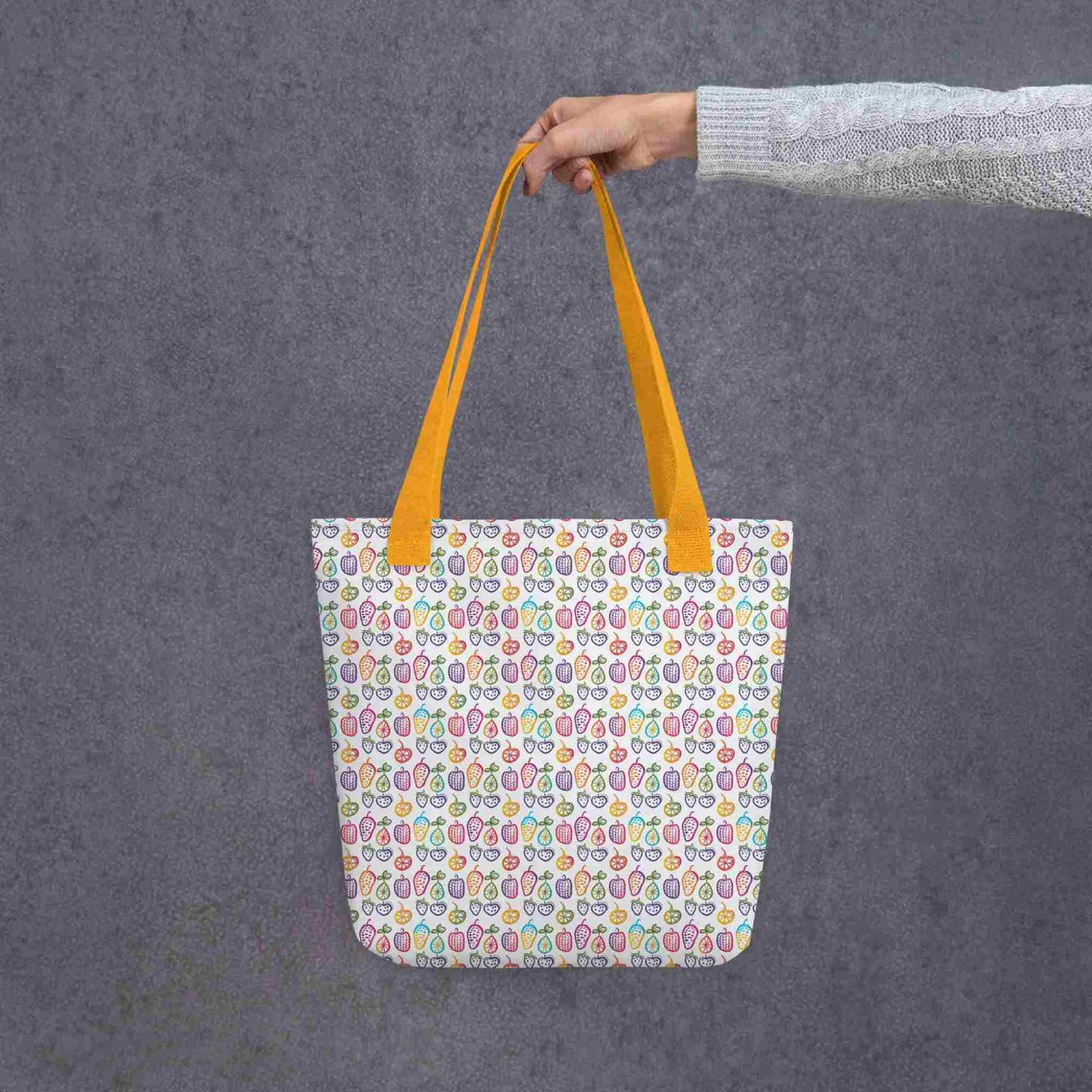 A fruit tote bag with yellow handles, which features a colourful cheerful doodles of fruits in a crisp white background, held in a lady's hand. Dimension 15x15 inches. Grey background.