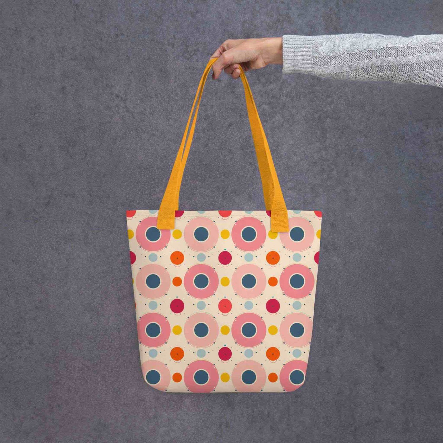 Artsy tote bag with yellow handles, which features an artsy design of pastel candy pops of color in a beige background, held by a lady's hand. Dimension 15x15 inches. Grey background.