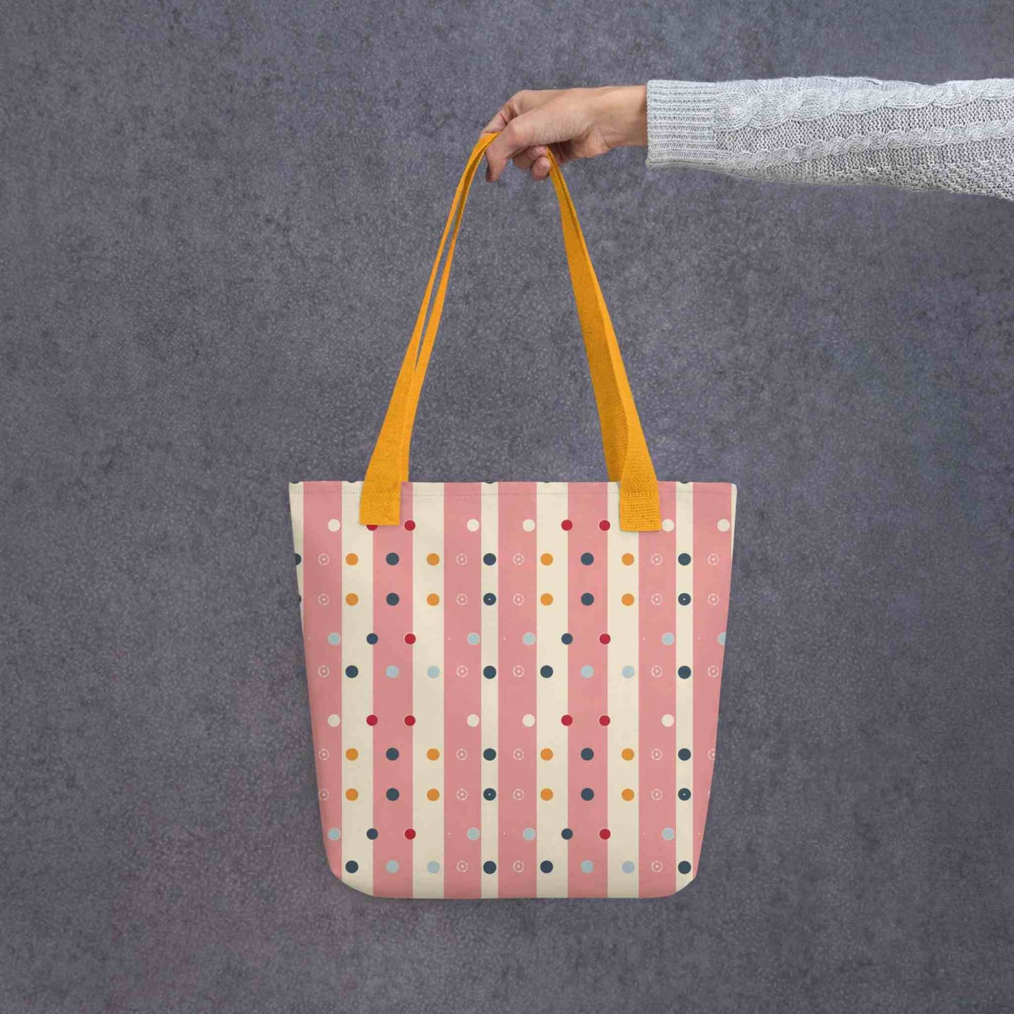 Pink striped tote bag with yellow handles, which features pink and beige stripes with colourful pokka dots, held by a lady's hand. Dimension 15x15 inches. Grey background.