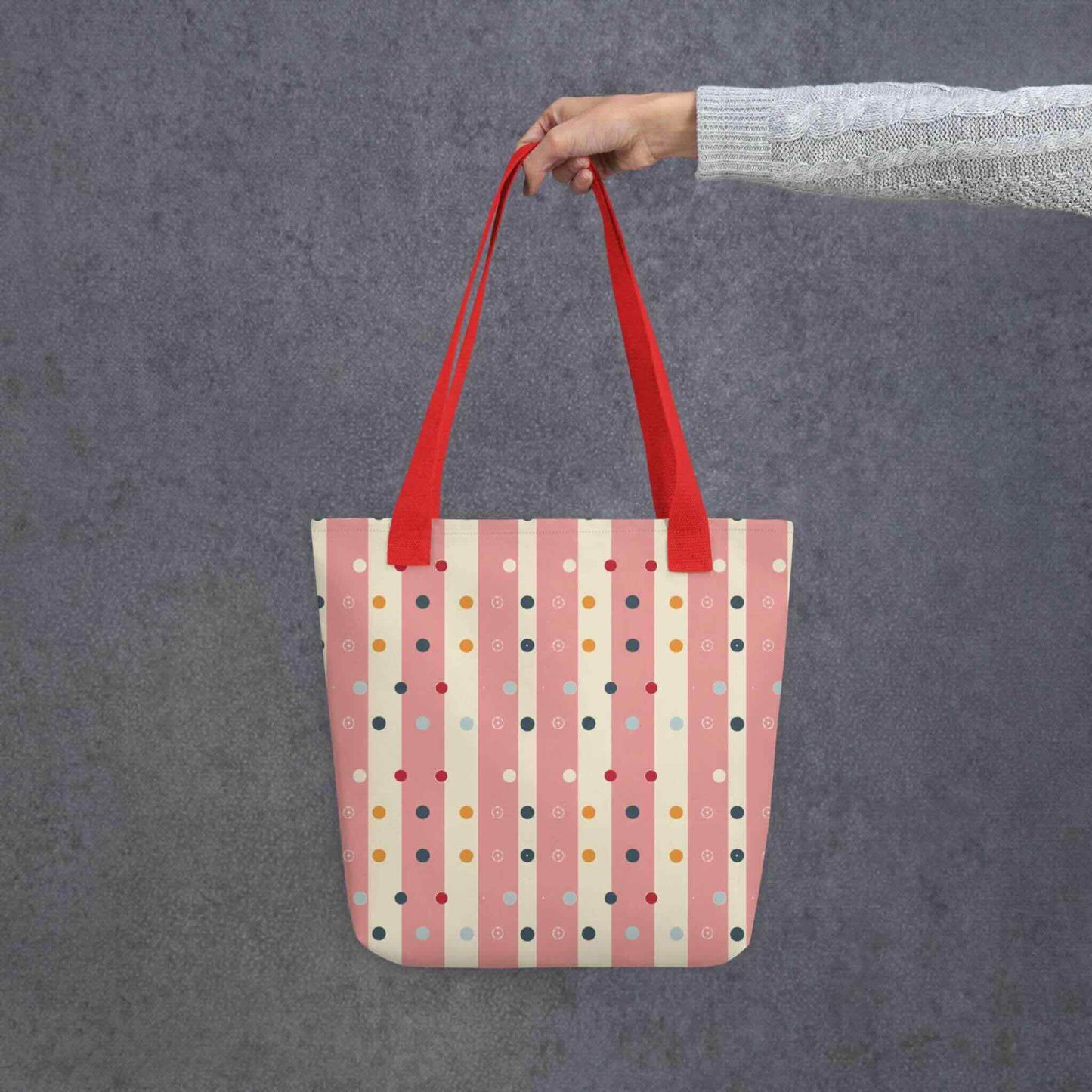 Pink striped tote bag with red handles, which features pink and beige stripes with colourful pokka dots, held by a lady's hand. Dimension 15x15 inches. Grey background.