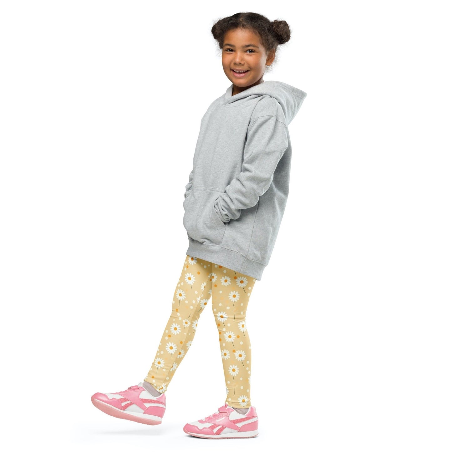 Front left view of a smiling black girl whose hair is bound into buns wearing a grey sweater, and leggings which feature sweet daisy prints on an egg yellow background. The girl is standing against a white background.