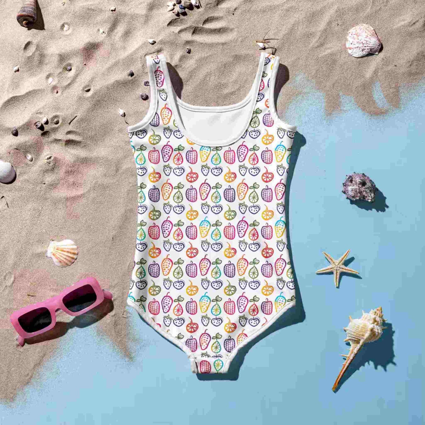 Flatlay of an adorable junior one-piece swimsuit which features a cheerful pattern of colorful fruit doodles across a white background., lying rear side up on a blue floor with sea sand. A set of pink sunglasses, pebbles, starfish and sea shells lie around the swimsuit.