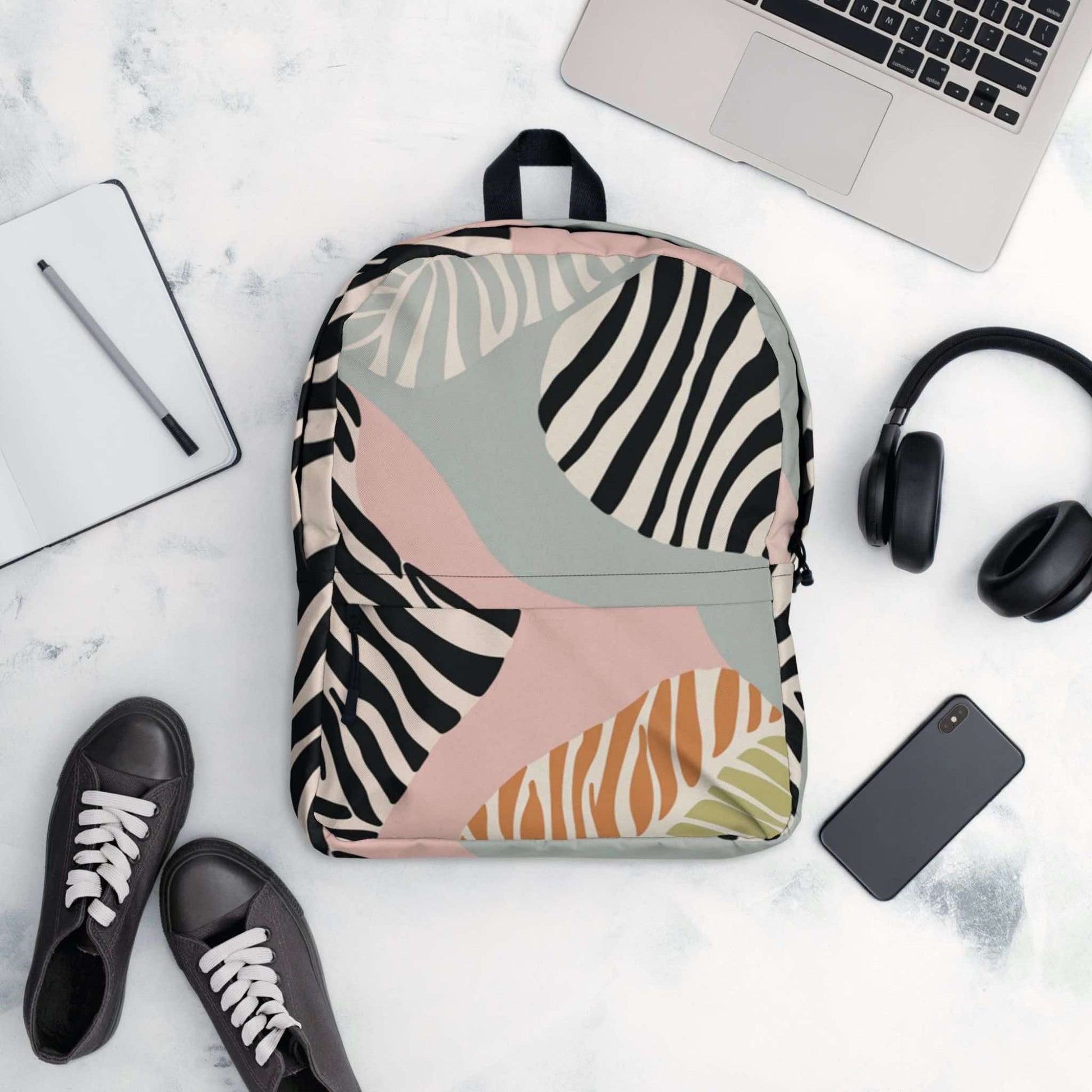 A flatlay of a zebra print backpack, surrounded by items it could conain, like a laptop, handphone, headphone, ontepad and pen, and even shoes.