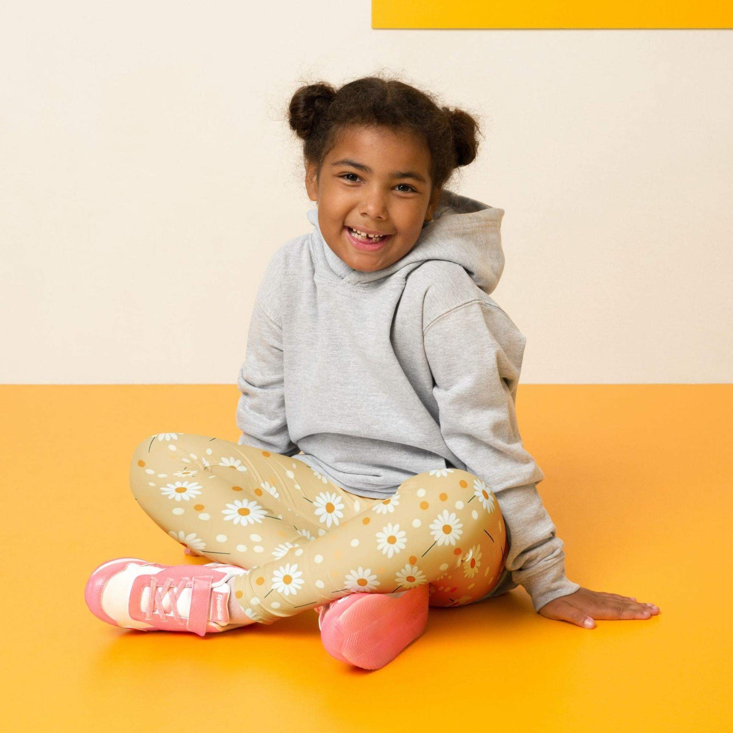 Front view of a smiling black girl whose hair is bound into buns wearing a grey sweater, and leggings which feature sweet daisy prints on an egg yellow background. The girl is sitting cross-legged on a yellow floor, against a white and yelllow wall behind her