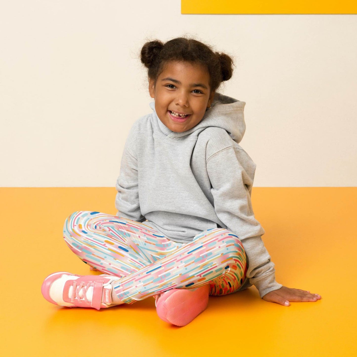 Front view of a smiling black girl whose hair is bound into buns wearing a grey sweater, and leggings which features playful candy colored lines in a white background. The girl is sitting cross-legged on a yellow floor, against a white and yellow wall behind her.