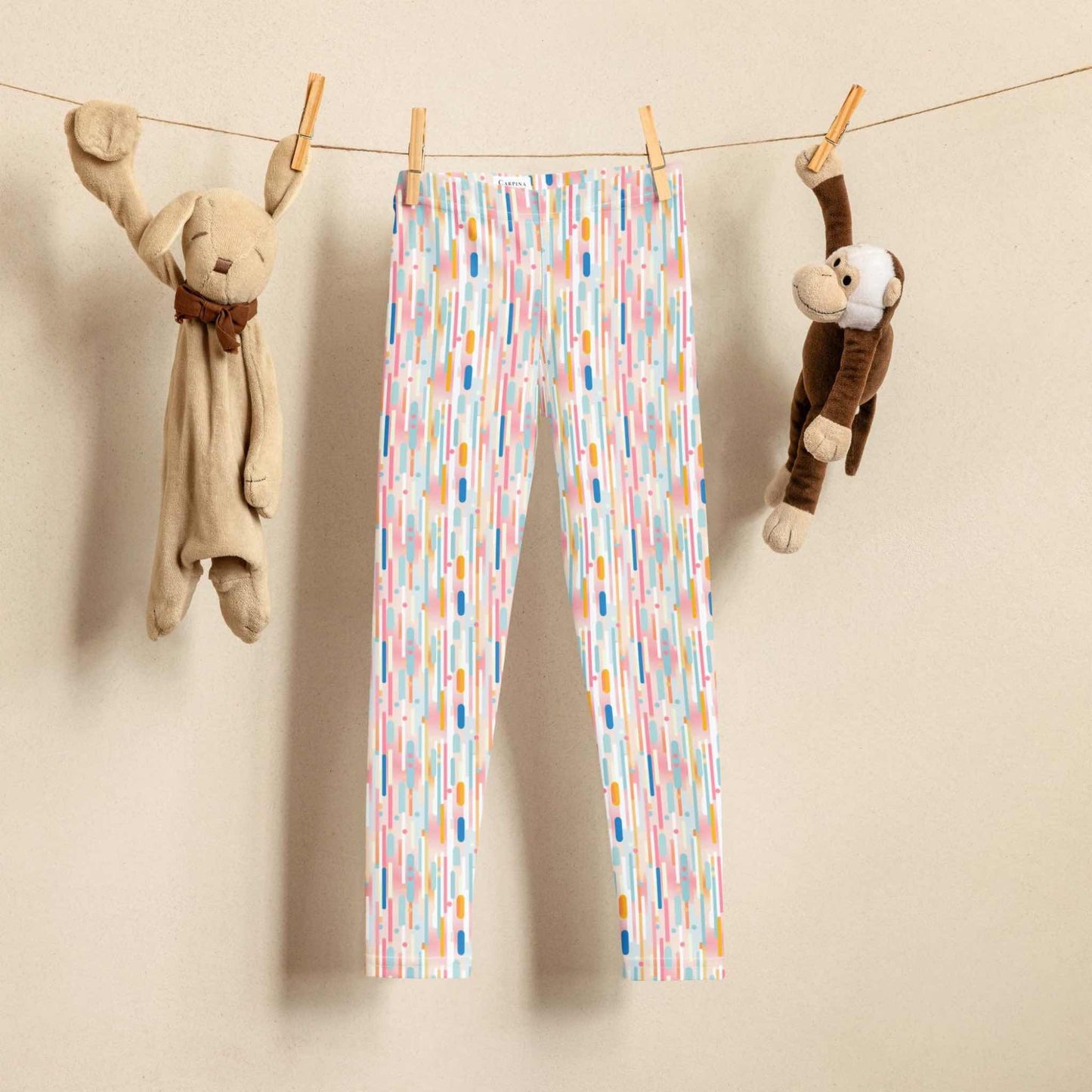 A pair of leggings which features playful candy colored lines in a white background is hung on a laundry line. A toy rabbit and toy monkey are hung on the same laundry line to the left and right of the leggings respectively. 