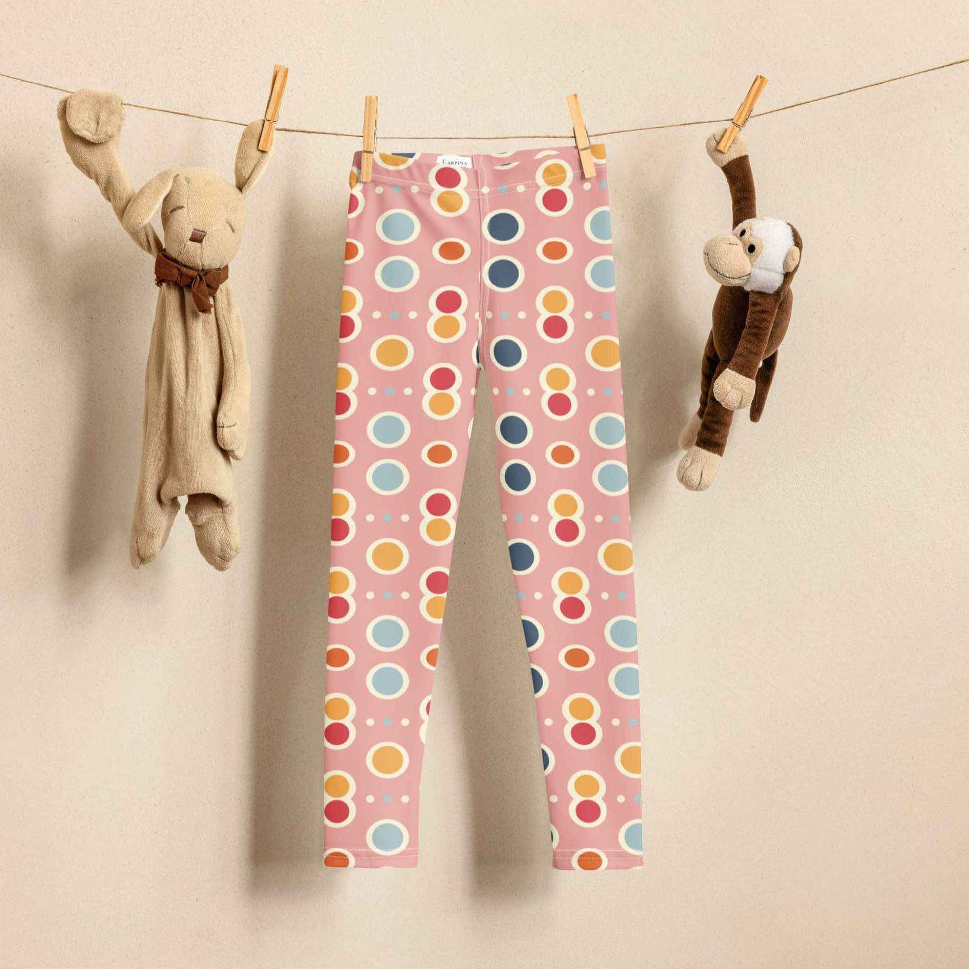 Pink leggings 2T and larger sizes! These adorable leggings feature a playful mix of polka dots and cute shapes in cheerful pastel colors on a sweet pink background, hanging on a laundry line, next to a toy bunny and toy monkey. Biege background.  