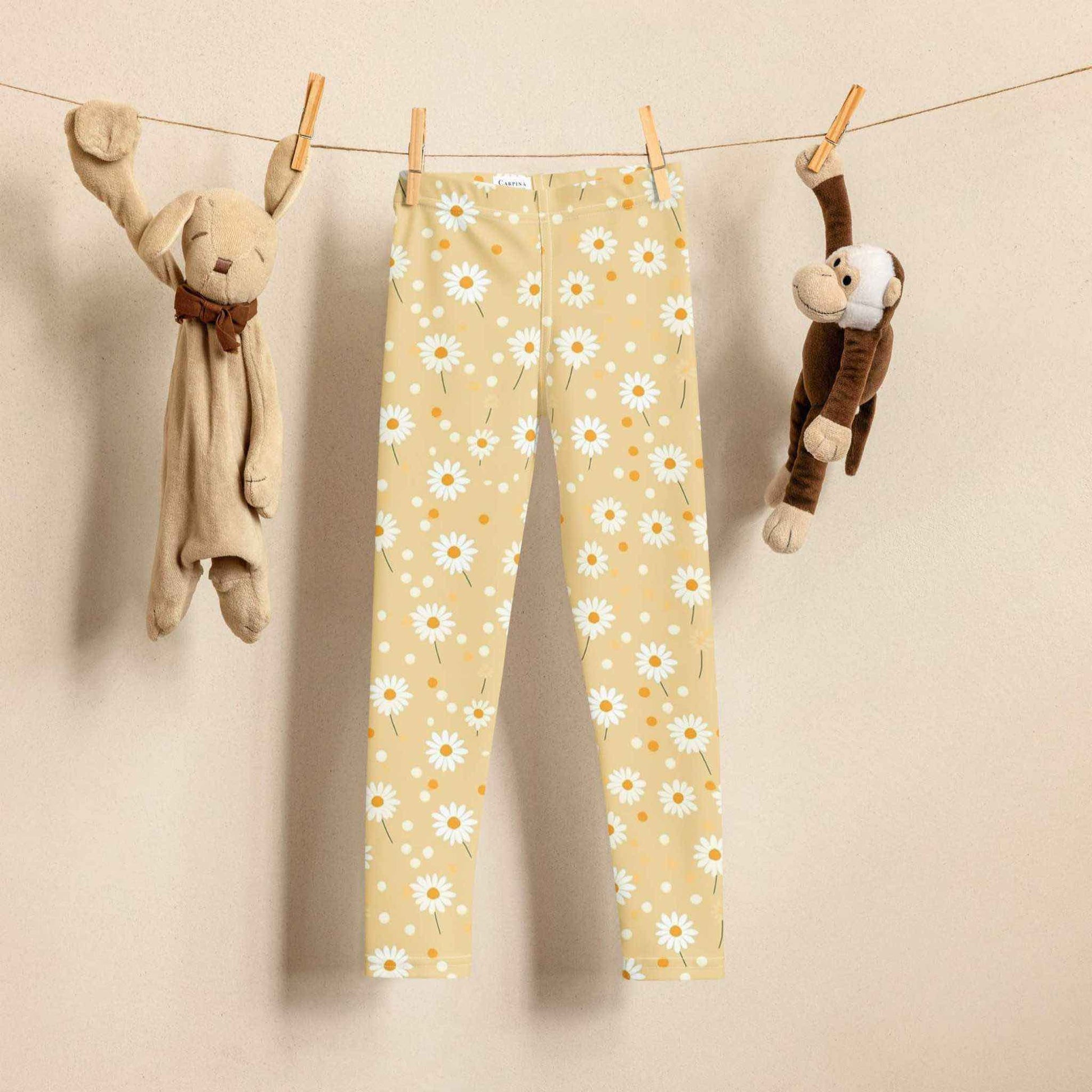 2T yellow leggings named DAISYDOODLE for toddlers, and bigger sizes for kids. This design features playful daisies &amp; polka dots on a bright egg yellow background, hanging on a clothes line next to a toy bunny and toy monkey. Biege background.