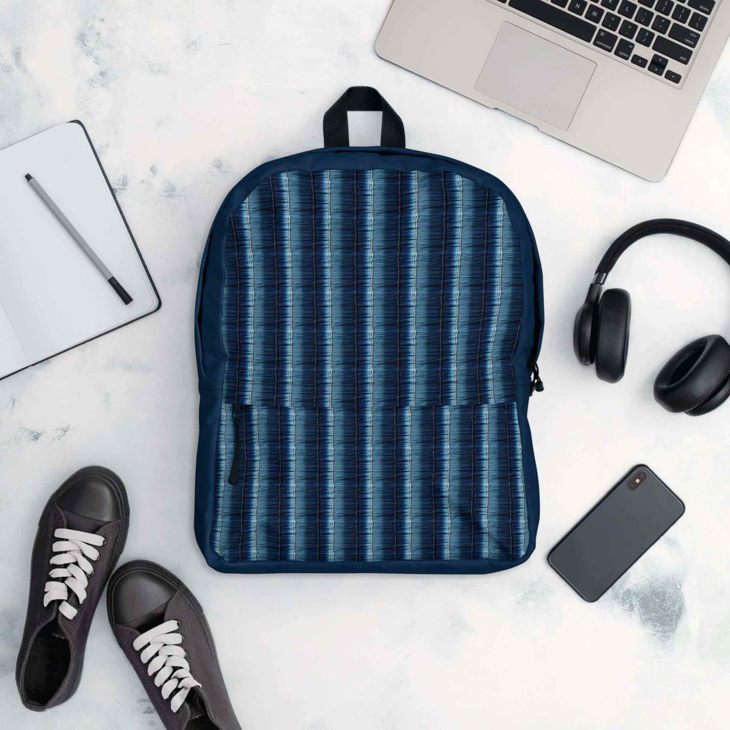 Flatlay of denim stripe backpack, facing up, with a notepad, shows, notebook computer, earphones and mobile phone lying next to it, against a white background.