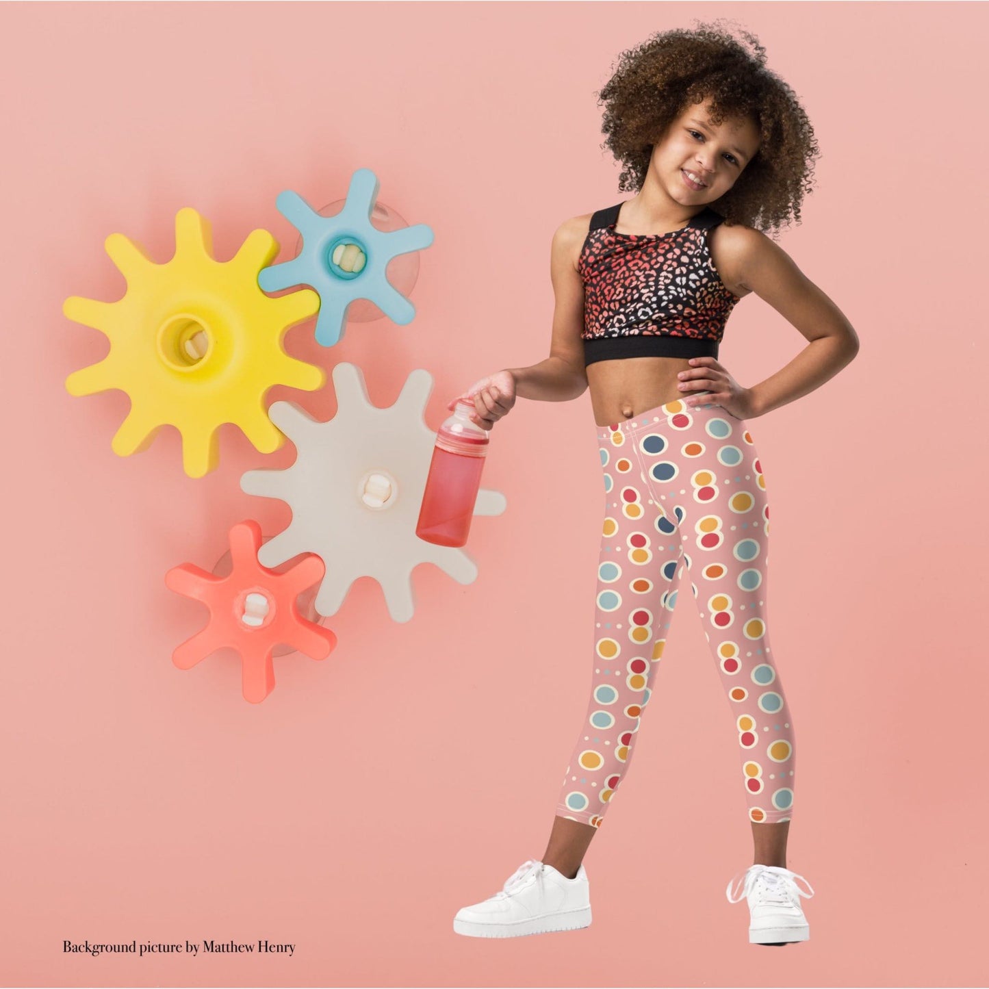 A black gril wearing a crop top and leggings which features a playful pattern of pastel and white circles on a pink background. The girl stands in a playful pose agaist a pink wall with colourful wheel cogs.