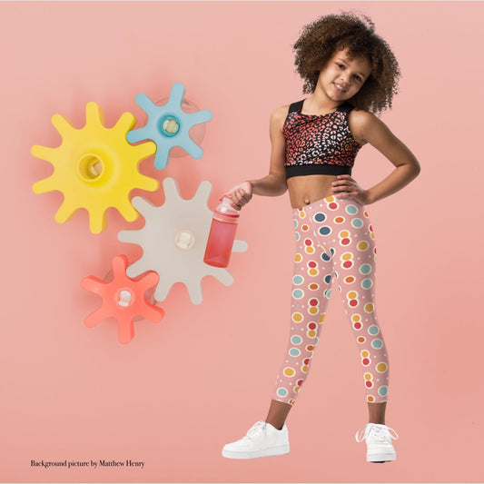 A black gril wearing a crop top and leggings which features a playful pattern of pastel and white circles on a pink background. The girl stands in a playful pose agaist a pink wall with colourful wheel cogs.