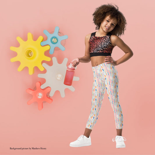 Kid and roddler leggings which features playful candy colored lines in a white background, worn by a young girl. The girl stands in a playful pose agaist a pink background with colourful cogs.
