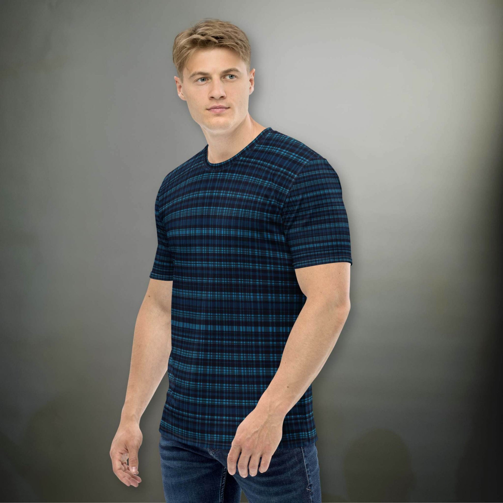Male Caucasion in a plaid dark blue all-over-print tshirt, in a plaid design that blends modern structure with timeless charm, offering a sophisticated yet versatile look. The man, in a pose where his head is tilted to his left and his body to his left, stands in front of an illuminated dark grey wall.