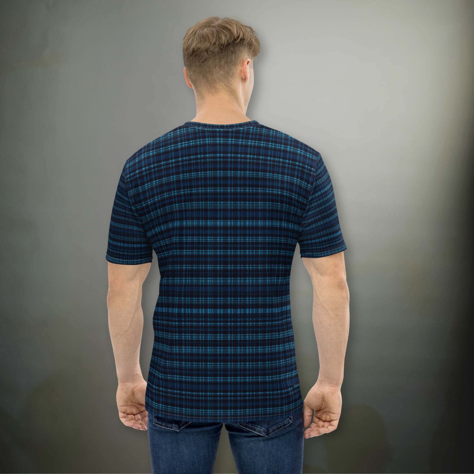 Male Caucasion in a plaid dark blue all-over-print tshirt, in a plaid design that blends modern structure with timeless charm, offering a sophisticated yet versatile look. The man, in a pose where his head is tilted to his left, stands in front of an illuminated dark grey wall.